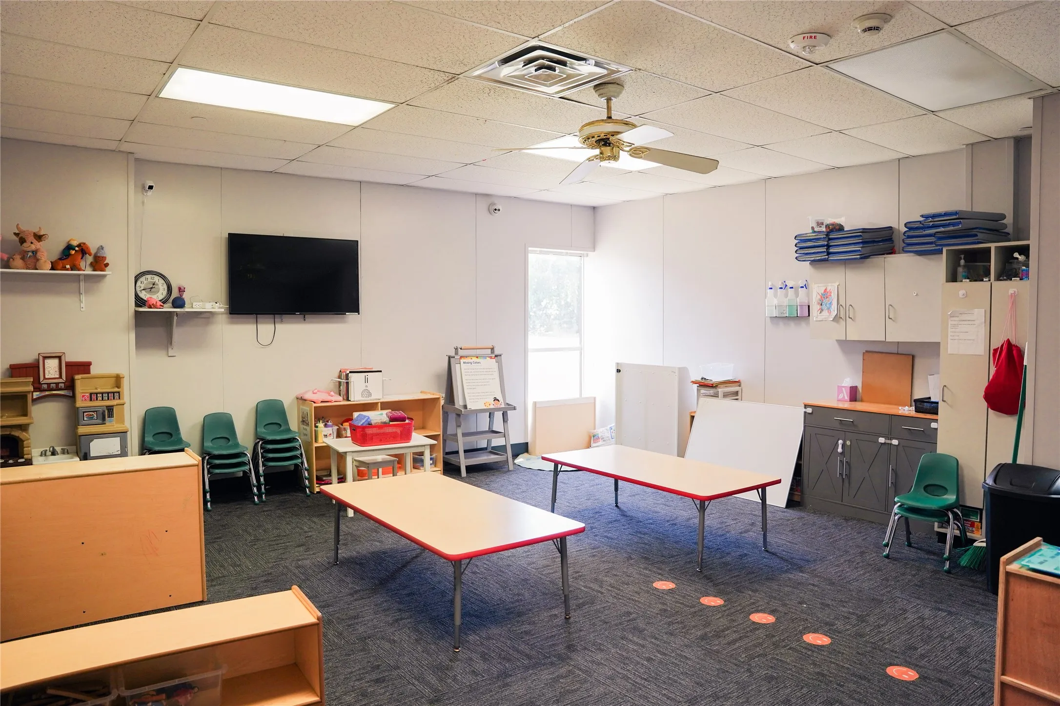 Versatile institutional or commercial space featuring durable carpet flooring, drop ceiling with integrated lighting and HVAC systems, multiple built-in storage shelving units, wall-mounted flat-screen display, and generous open floor plan with ample natural light from windows and glass entry door