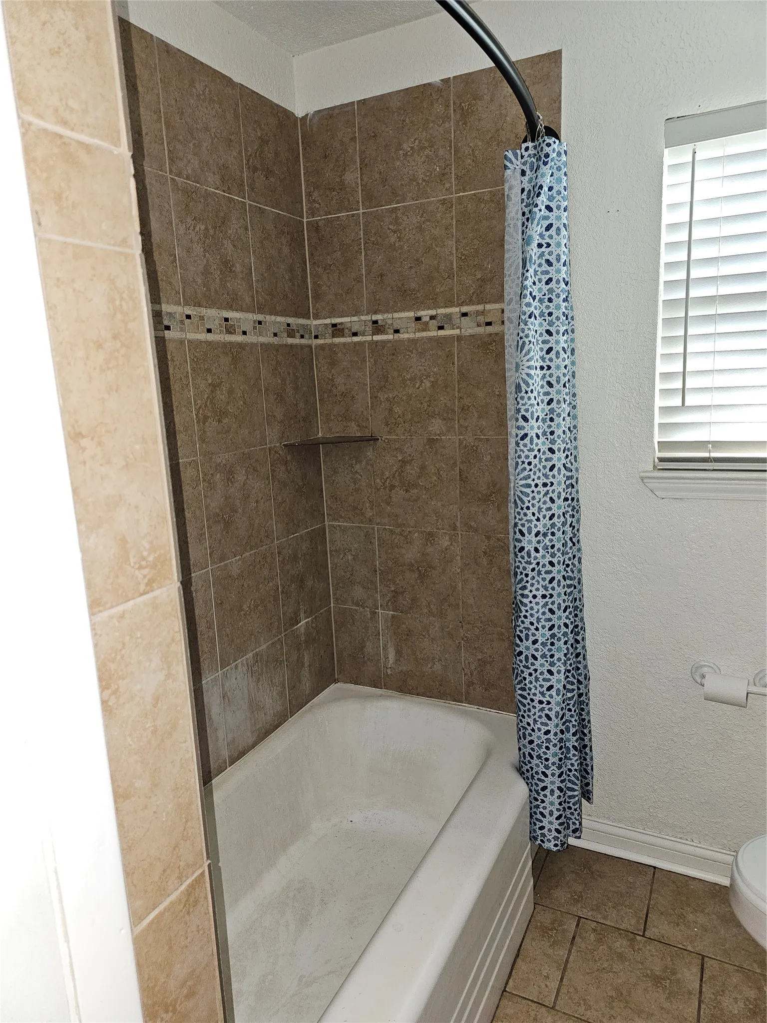Spa-inspired bathroom shower featuring rich earth-tone ceramic tile surround, decorative mosaic accent band, built-in shelf niche, and curved glass enclosure with coordinating patterned curtain liner