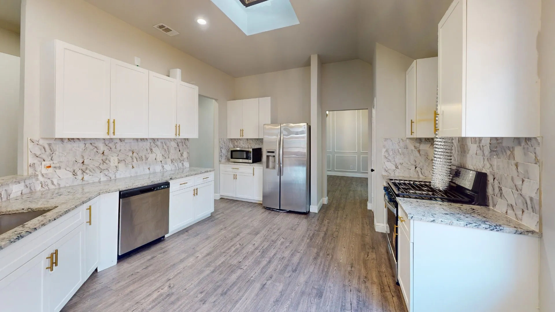 Galley-style kitchen featuring white cabinetry with brushed gold hardware, marble subway tile backsplash, stainless steel appliances including a gas range and refrigerator, quartz countertops, wood-finish flooring, and skylight overhead for natural illumination
