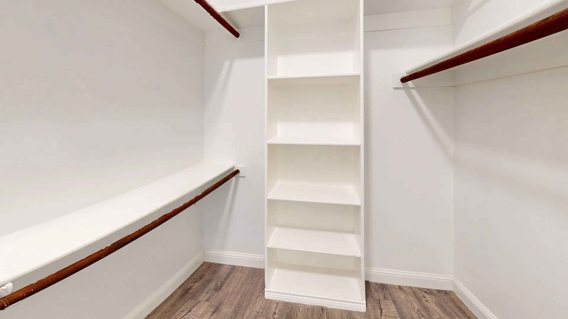 Spacious walk-in closet featuring dual clothing rods with ample hanging space, a central shelving unit with multiple compartments for organized storage, and wood-finish flooring throughout