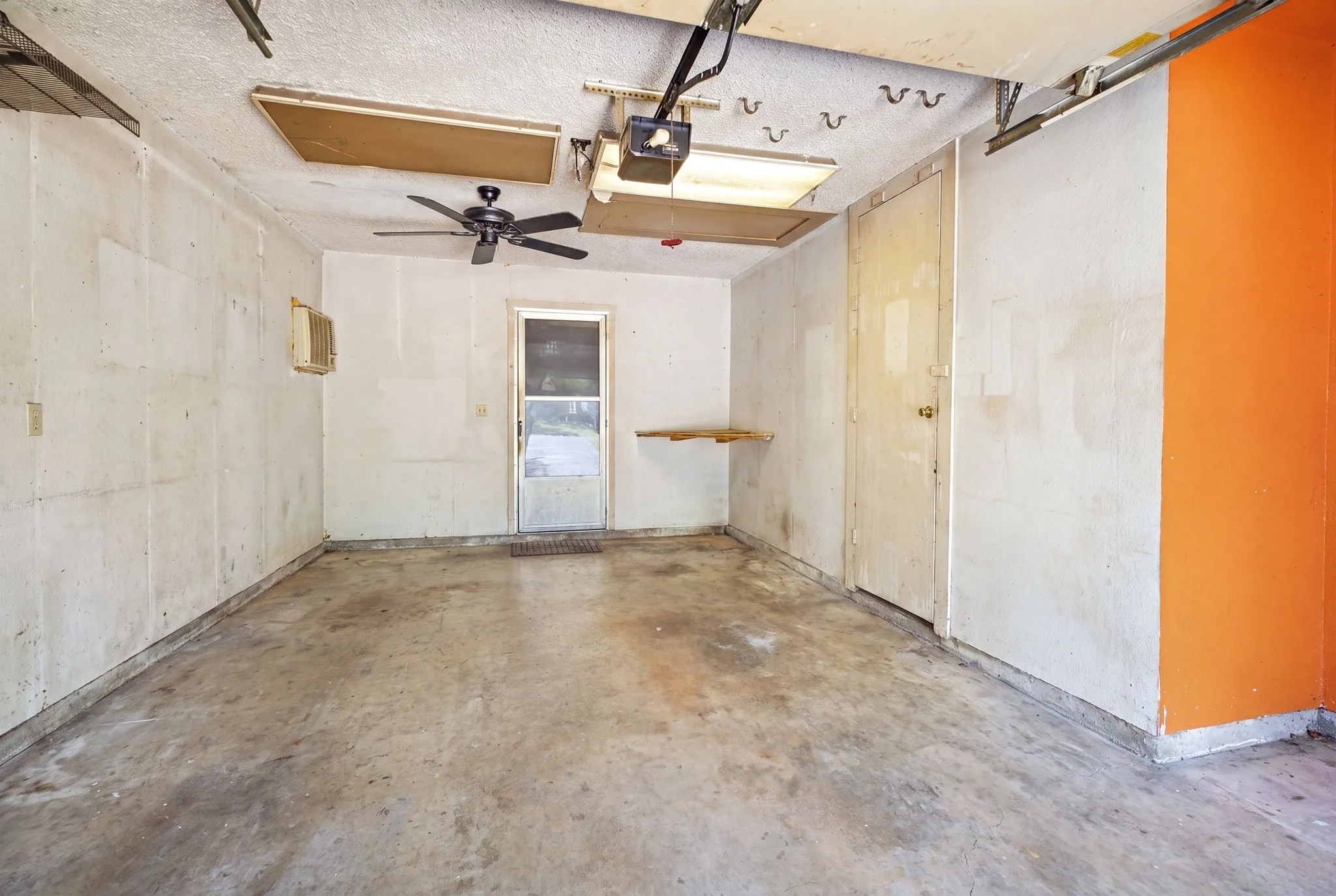 Interior garage space showing concrete floors, exposed ceiling rafters, ceiling fan, and accent wall.