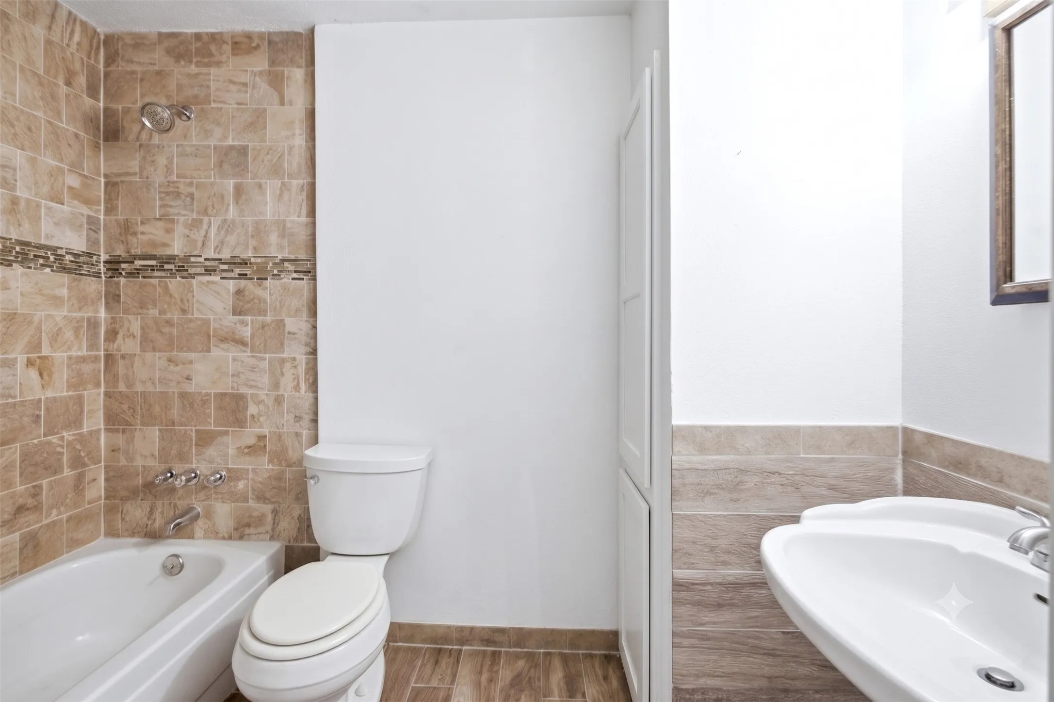Bathroom view showing a white toilet, tiled tub/shower wall, light wood flooring, and pedestal sink edge.