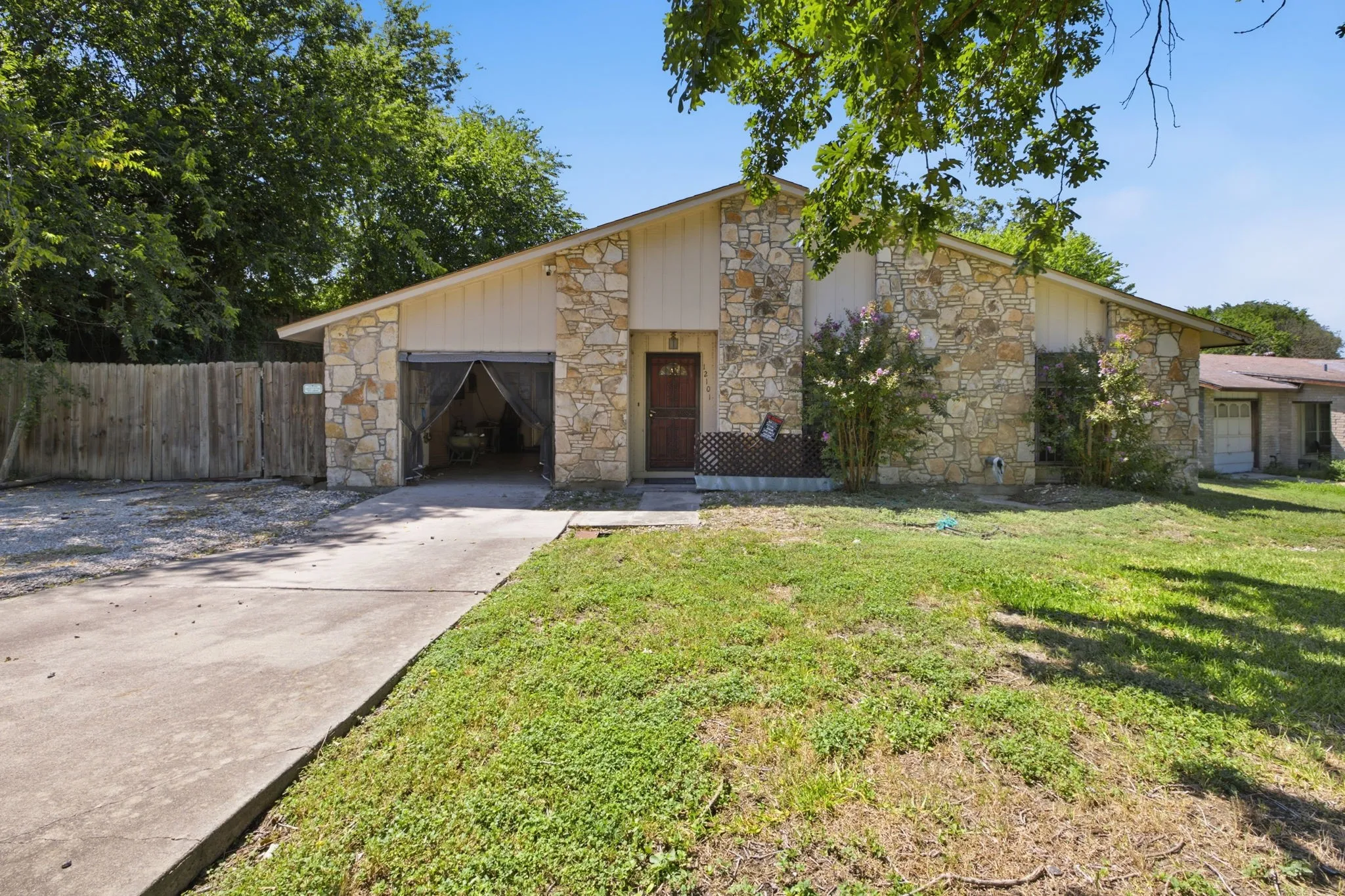 Daylight view of the stone exterior, driveway leading to the garage, and surrounding green grass and trees.
