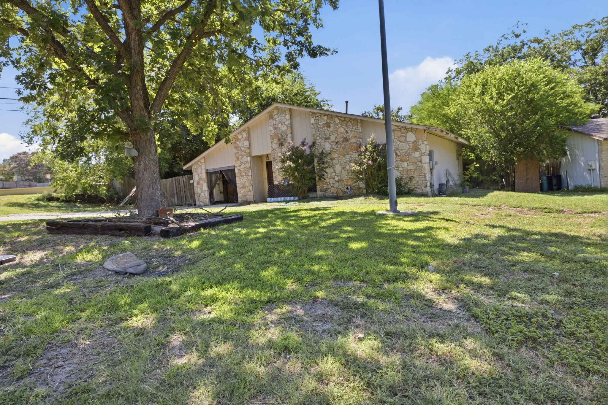Front yard view featuring a well-manicured lawn, mature trees, and a side view of the single-story stone house.