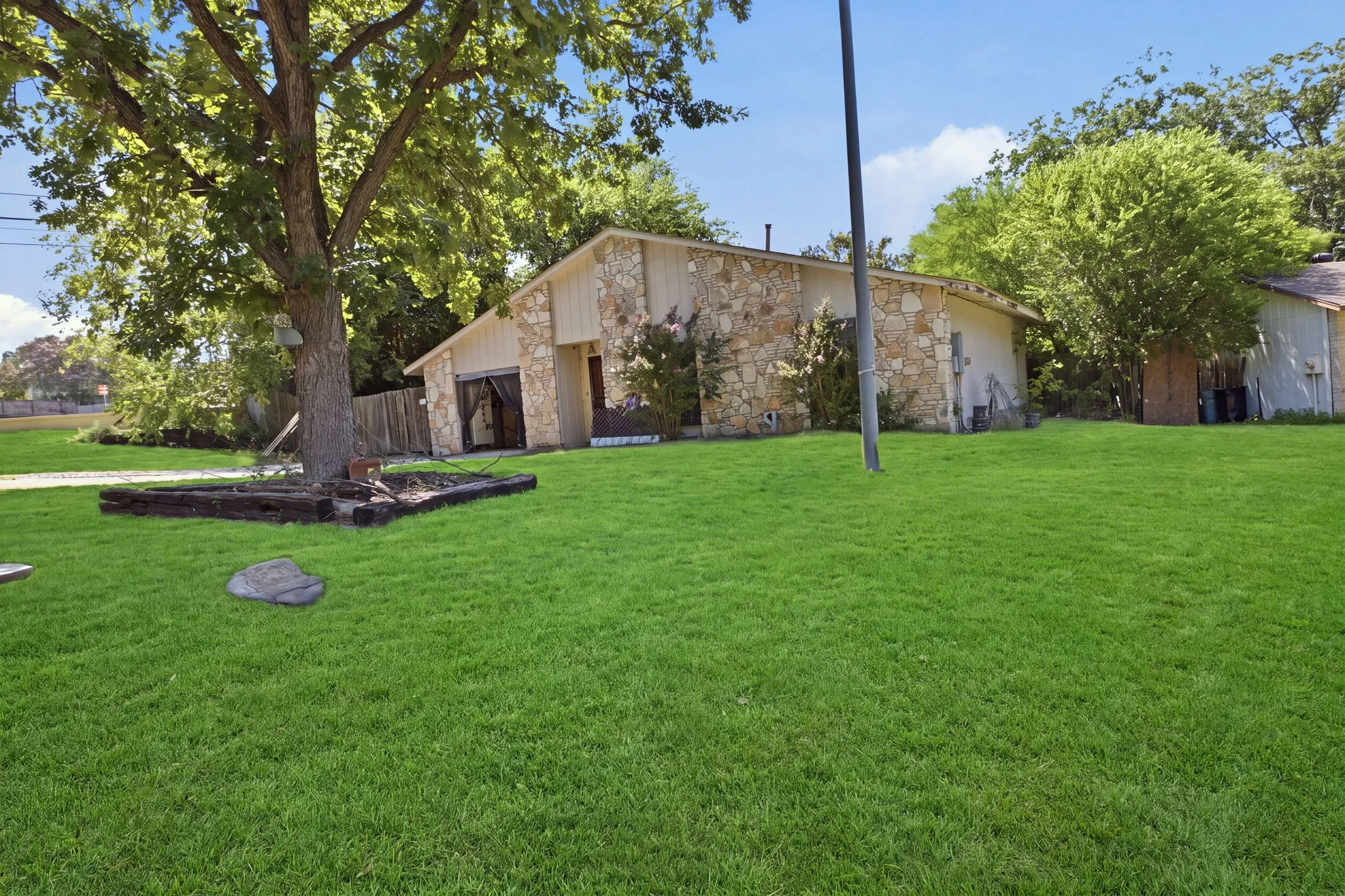 Wide front view showing a lush green lawn, mature shade trees, and the stone home in the background.