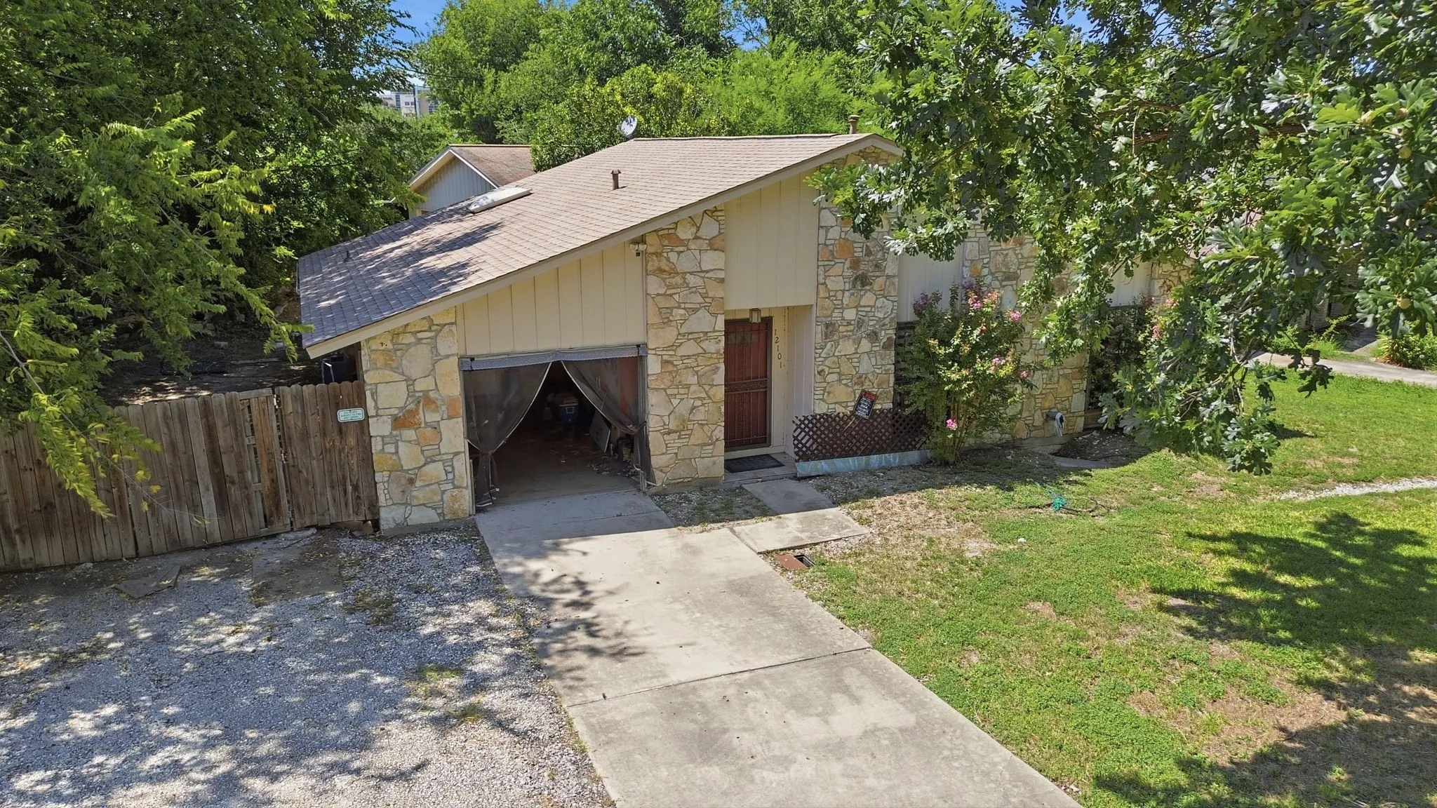 Elevated front shot of the stone home facade, garage, paved driveway, and surrounding shade trees.