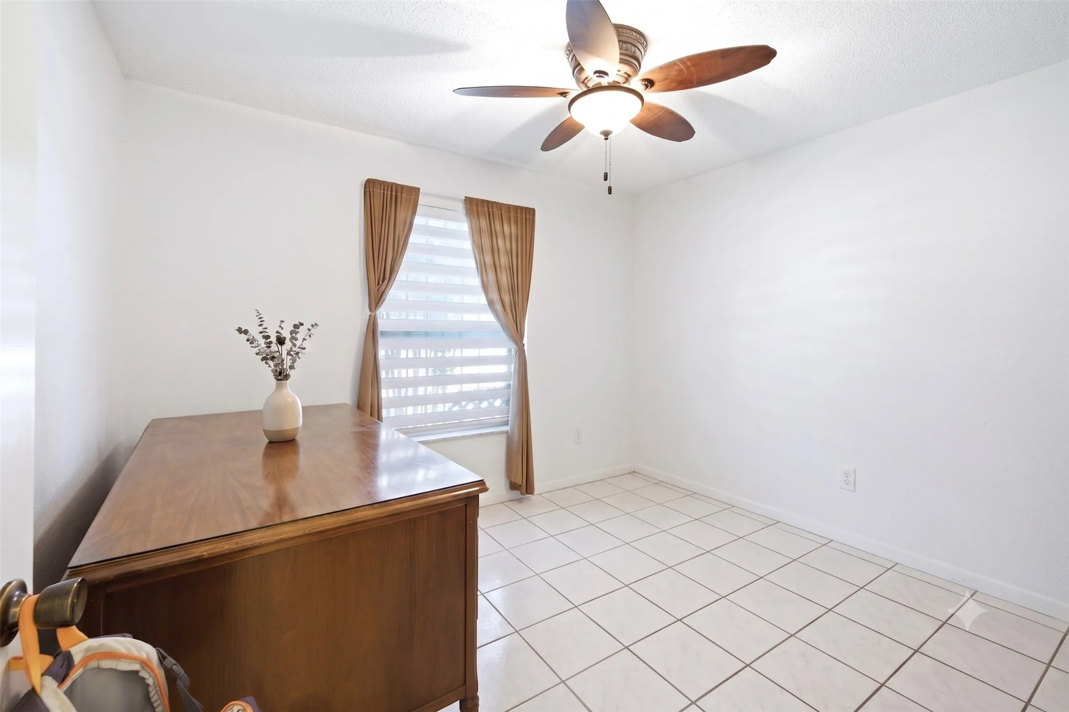 Bright office space with tiled floors, a ceiling fan, a window with curtains, and a wooden desk.