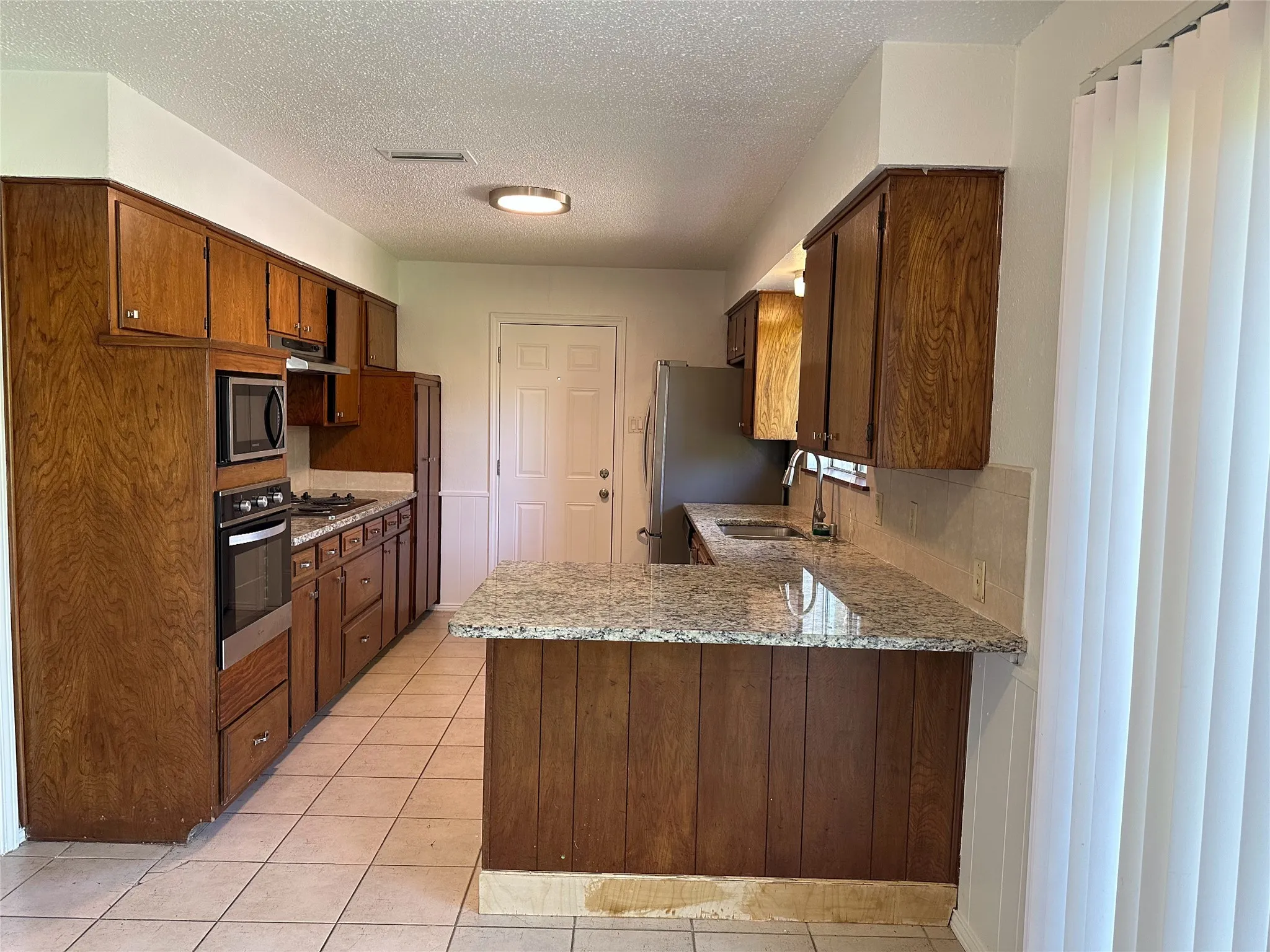 Kitchen featuring wood cabinetry, a built-in oven and microwave, stone countertops, a tile backsplash, and tile flooring