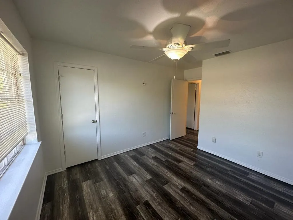 Spacious bedroom featuring dark wood-finish flooring, ceiling fan with integrated lighting, and abundant natural light from large window