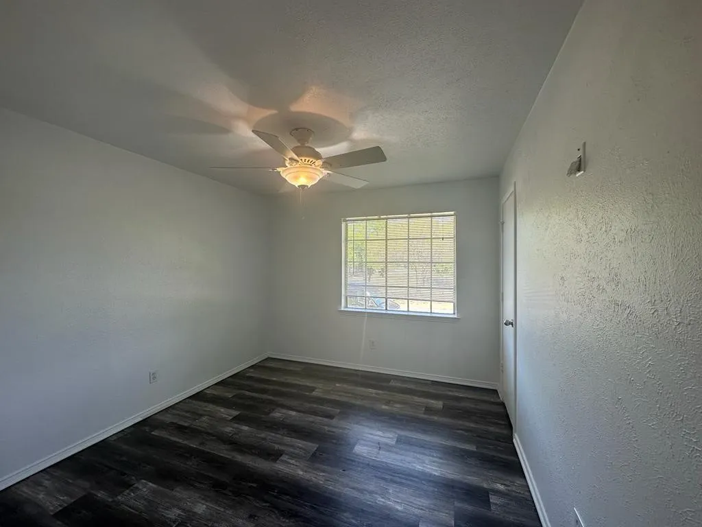 Spacious bedroom with dark wood-finish flooring, ceiling fan with integrated lighting, and natural light from large gridded window