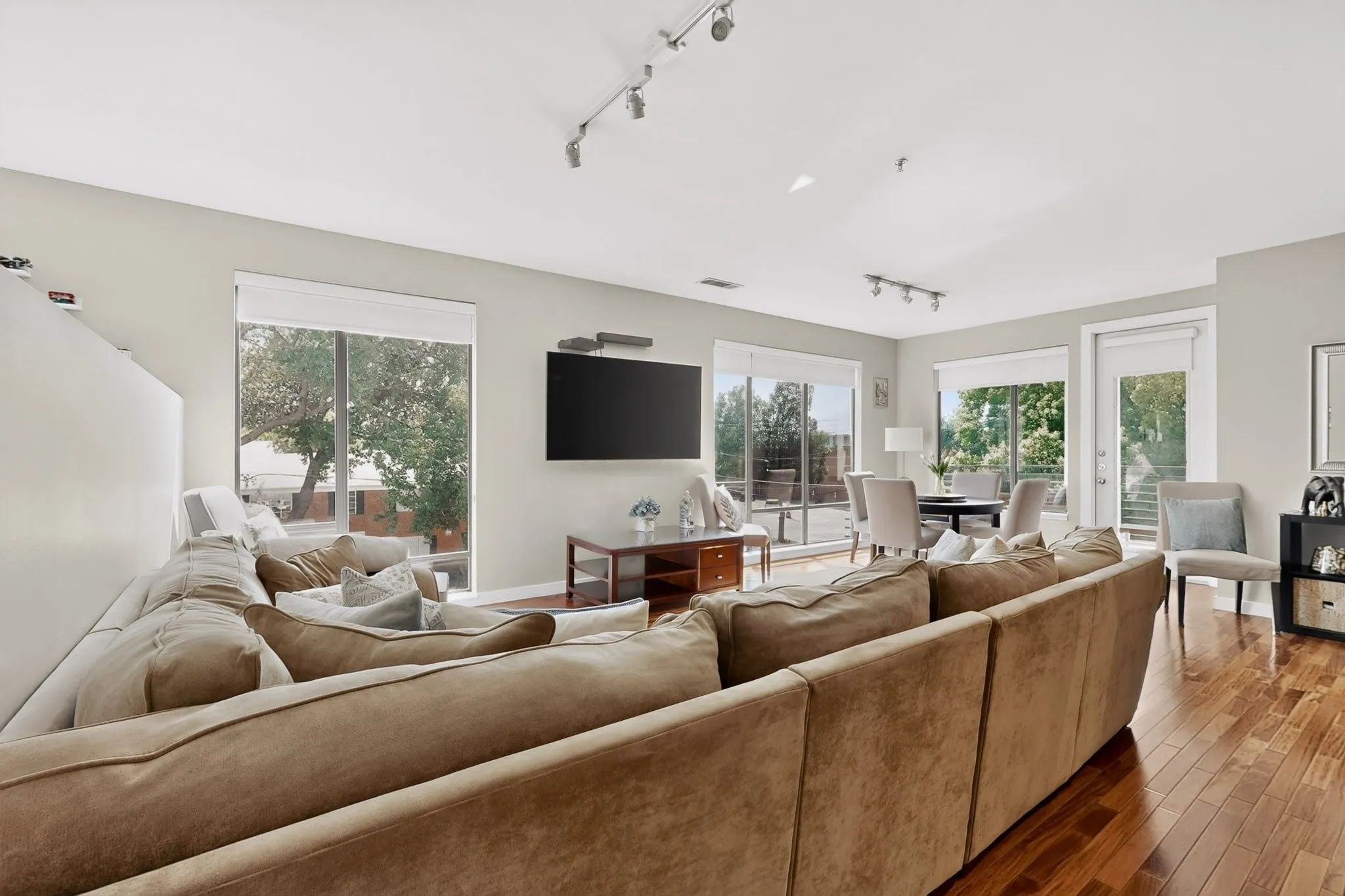 Spacious living area featuring rich wood-finish flooring, abundant natural light from large windows, and a glass door leading to an outdoor space