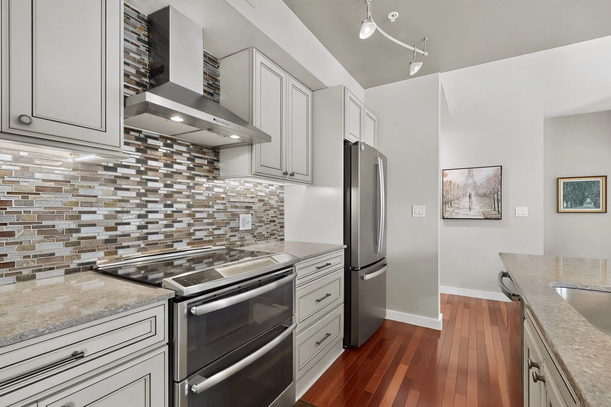 Kitchen featuring light-toned cabinetry with dark hardware, a stainless steel range hood, a mosaic tile backsplash, stainless steel appliances, and wood-finish flooring