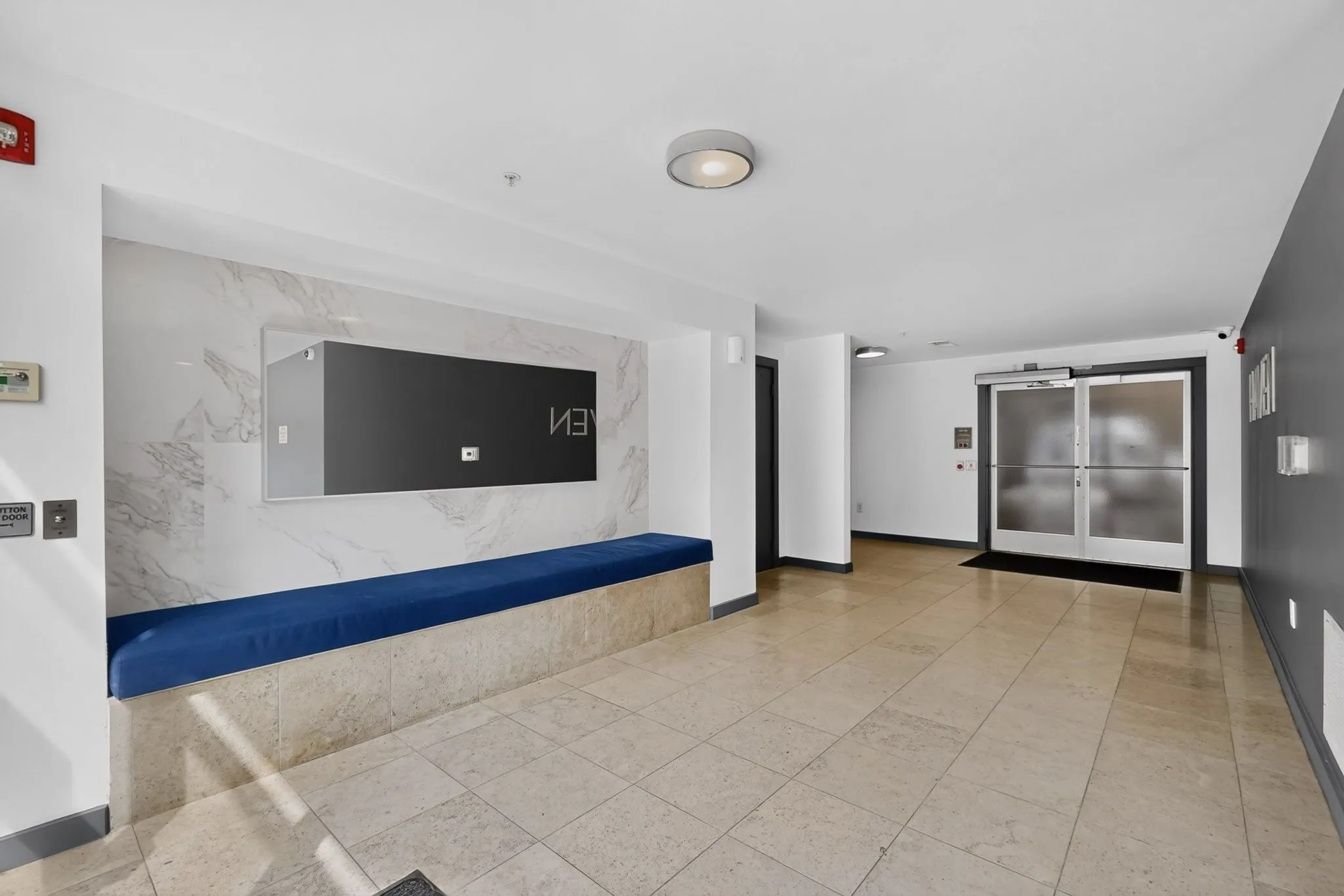 Lobby area featuring large format tile flooring, a built-in bench with blue upholstery, a wall-mounted display, and a frosted glass door entry