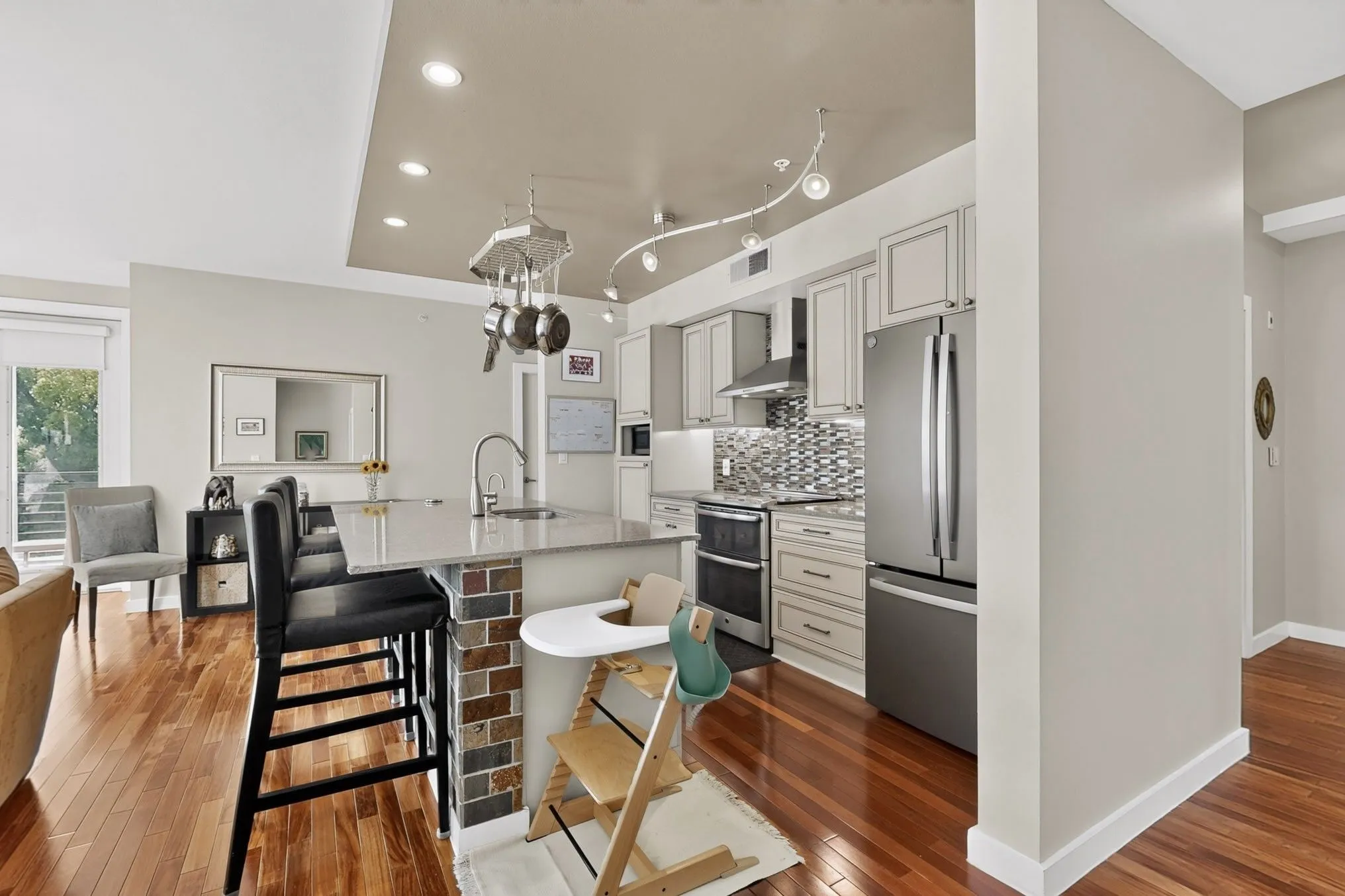 Contemporary kitchen featuring wood-finish flooring, light grey cabinetry, stainless steel appliances, a grey tile backsplash, and an island with a breakfast bar and brick-faced base