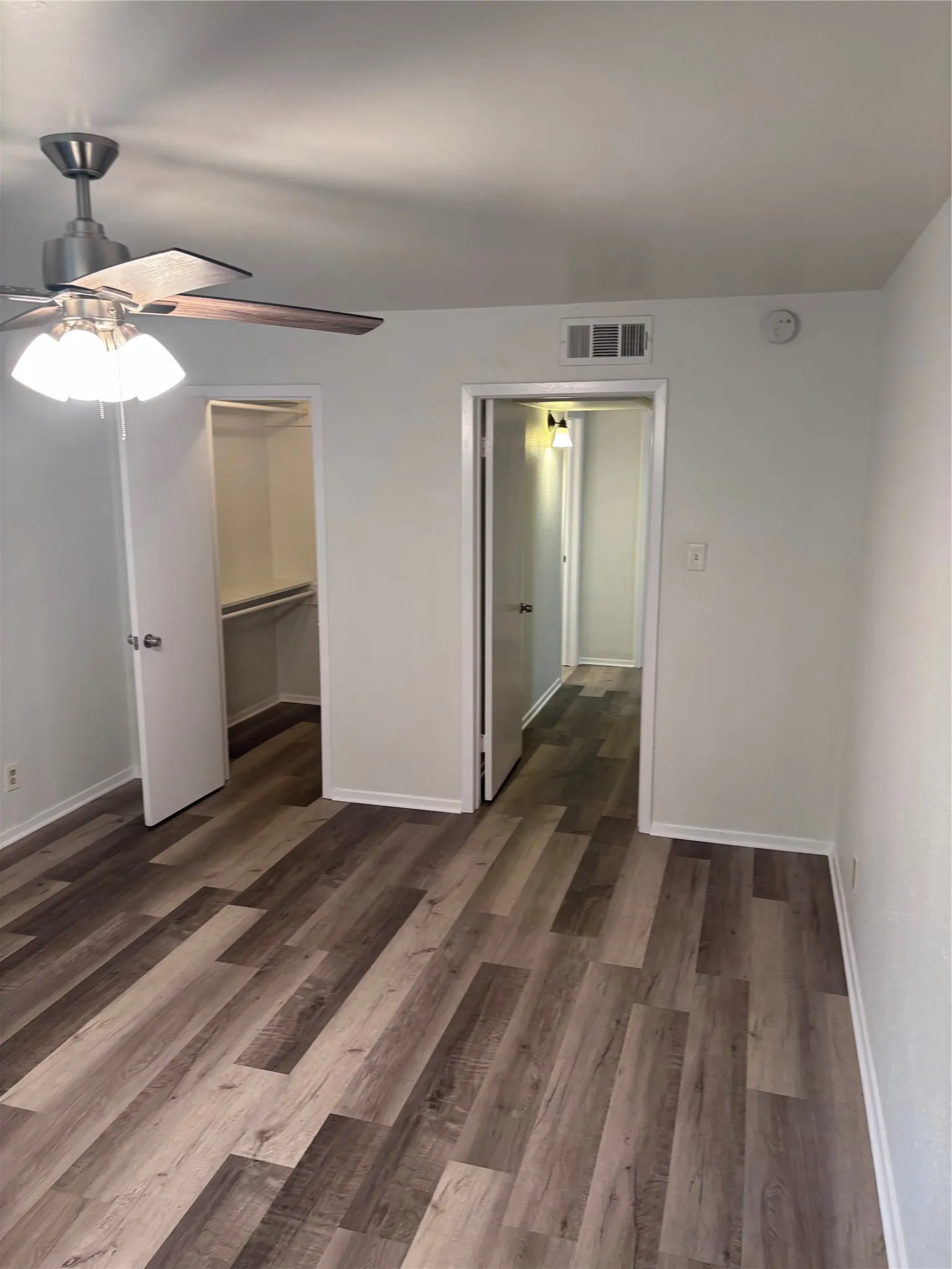 Spacious room featuring wood-finish flooring, a ceiling fan with integrated lighting, a built-in closet with shelving, and crisp white walls
