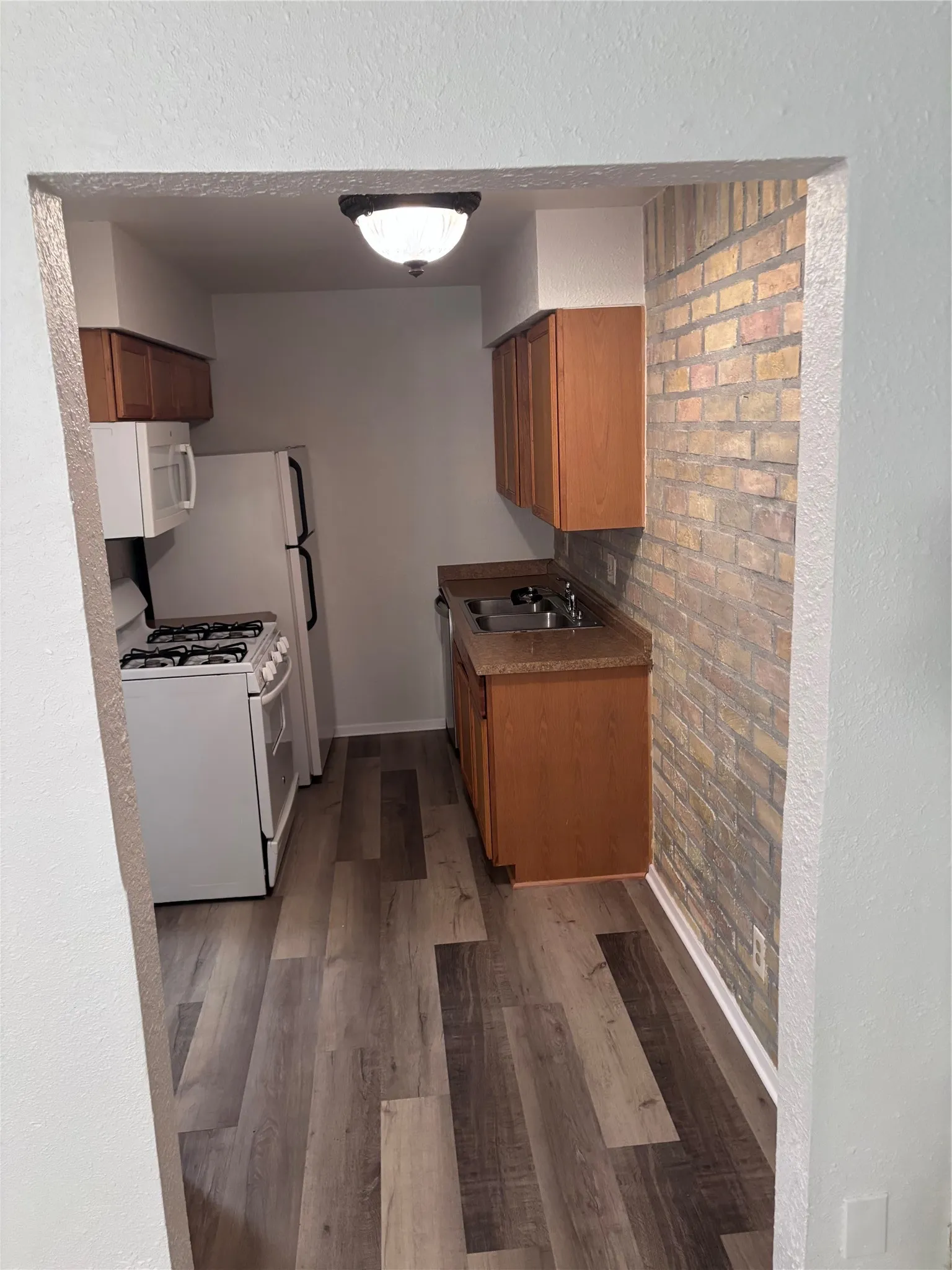 Galley kitchen featuring wood-finish flooring, gas range with stovetop, stainless steel sink with faucet, wood cabinetry, and brick accent wall