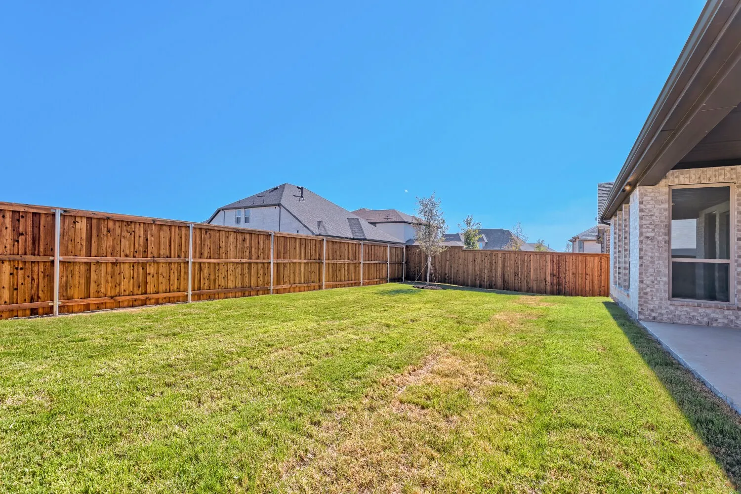 Expansive grassy backyard featuring a full privacy fence, covered patio, and brick exterior