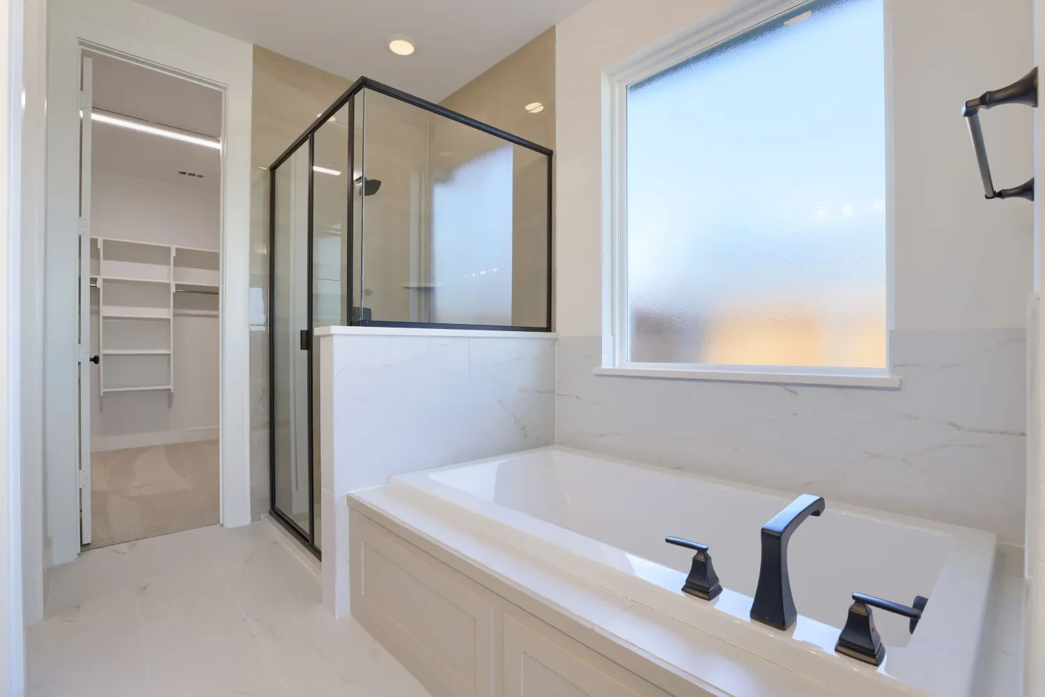 Freestanding soaking tub with dark-toned fixtures, walk-in shower with clear glass enclosure and dark-toned frame, light-toned wall tile, and built-in closet shelving