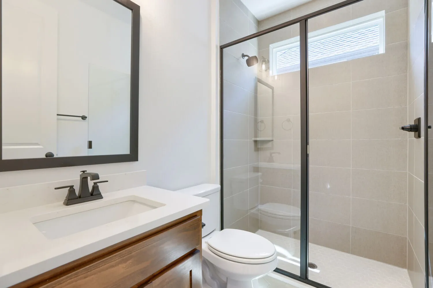 Modern primary bathroom featuring a floating wood-finish vanity with a white countertop and integrated sink, matte black faucet hardware, and a framed mirror
