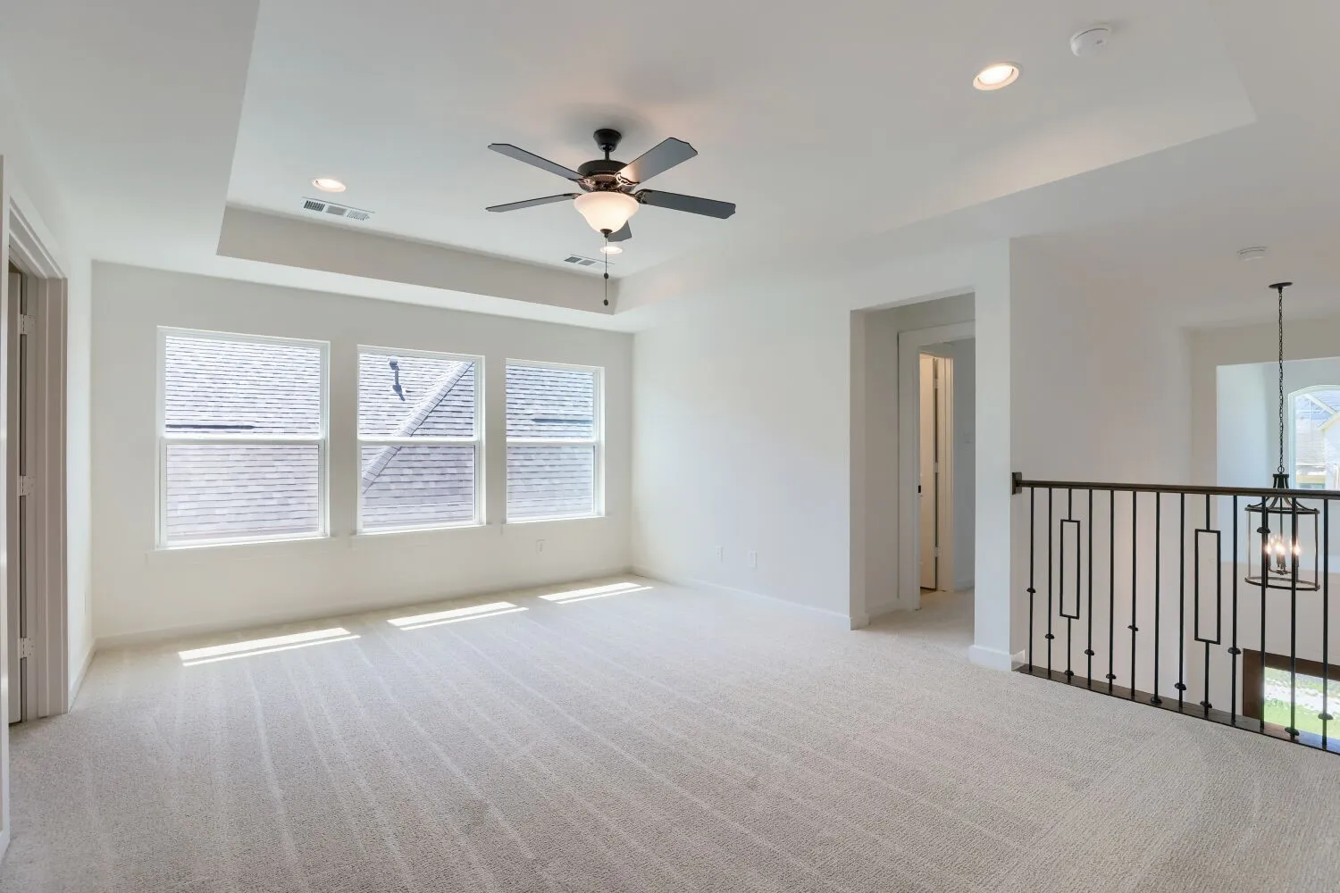 Bright and spacious primary suite featuring three large windows with plantation shutters, neutral carpet flooring, ceiling fan with integrated lighting, and direct access to a secondary balcony with metal railing