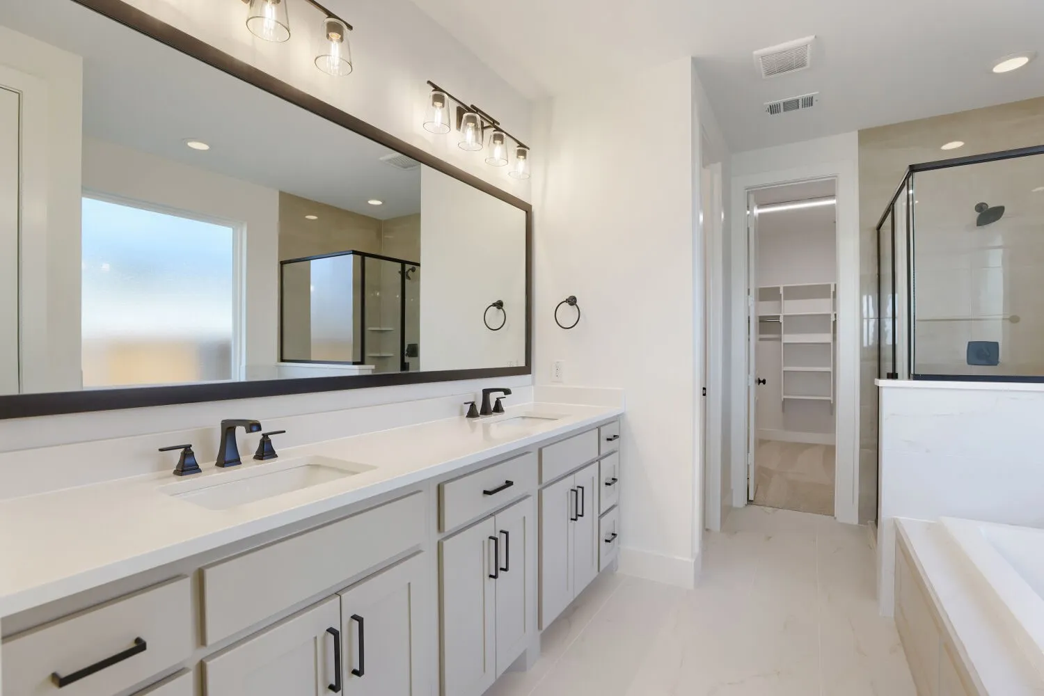 Primary Bathroom featuring dual vanity with white cabinetry, quartz countertops, and matte black hardware; large framed mirror with integrated lighting; walk-in shower with glass enclosure and tile surround; freestanding soaking tub; and recessed ceiling lighting throughout