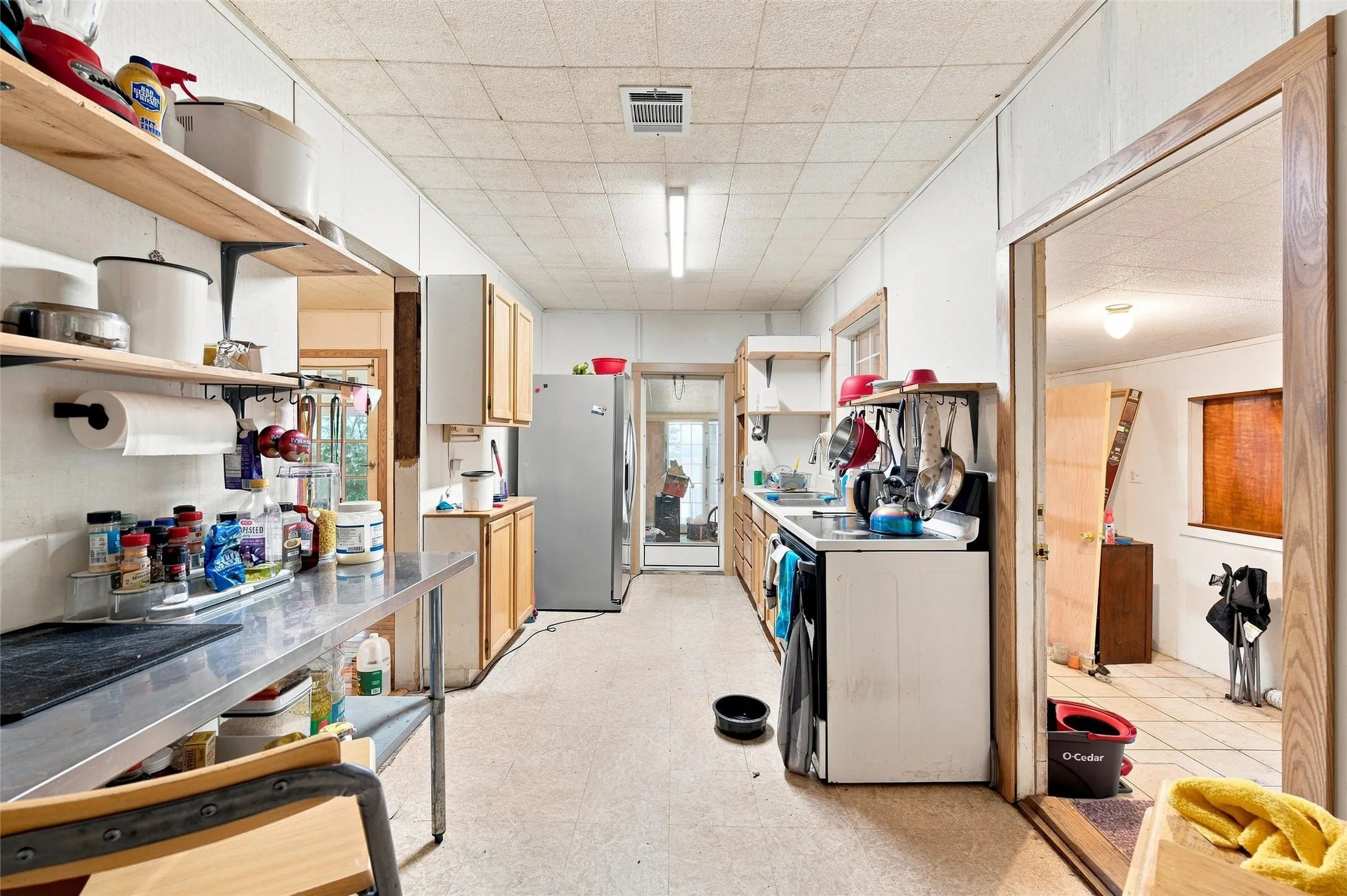 Galley-style kitchen featuring wood-finish cabinetry, stainless steel appliances including refrigerator and range, laminate countertops, open shelving with storage, and tile flooring throughout