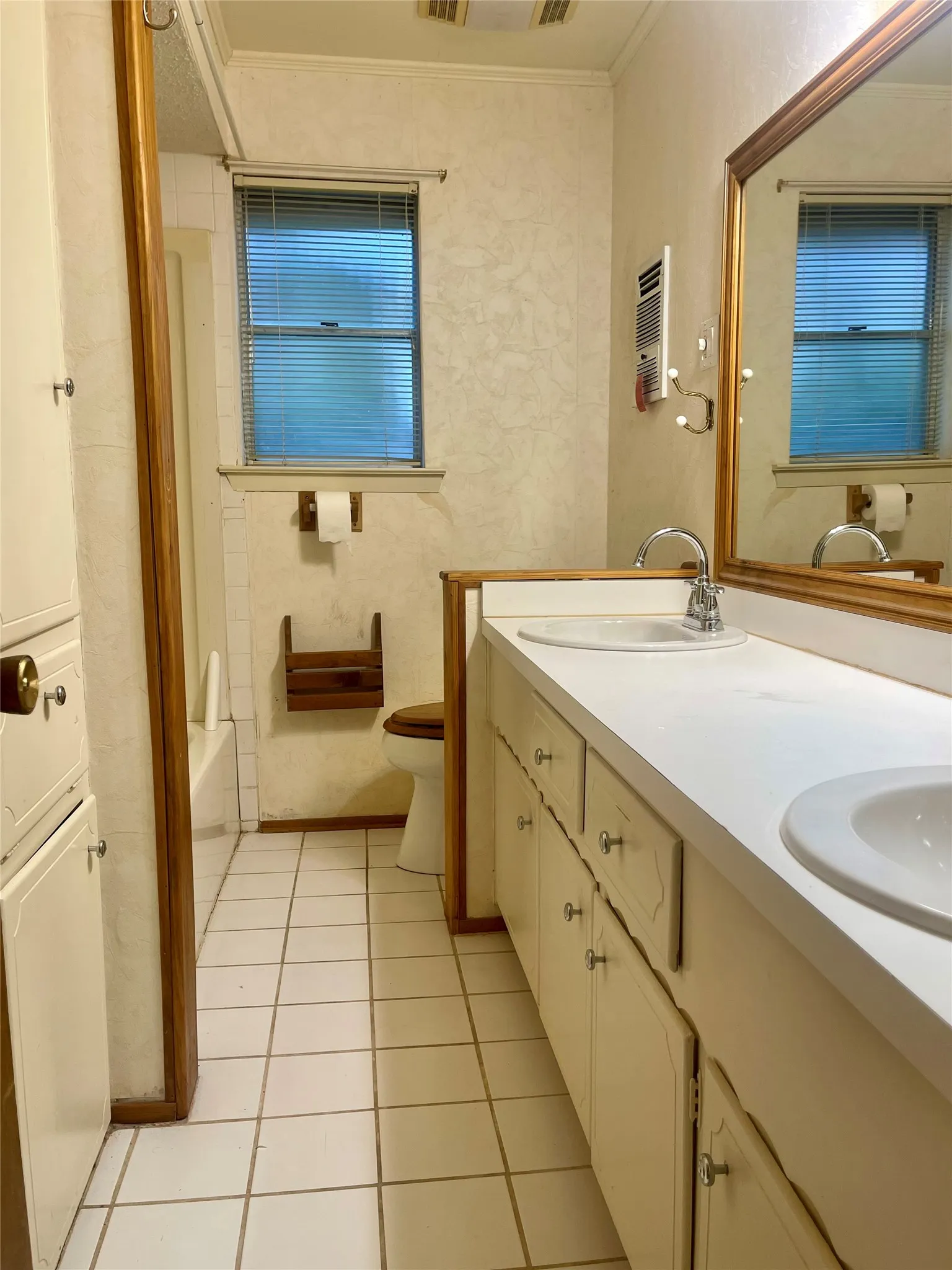 Galley-style bathroom featuring dual vanity with white countertop and integrated sink, wood-finish cabinetry with hardware pulls, large framed mirror, tile flooring, and operable window with blinds for natural light and ventilation