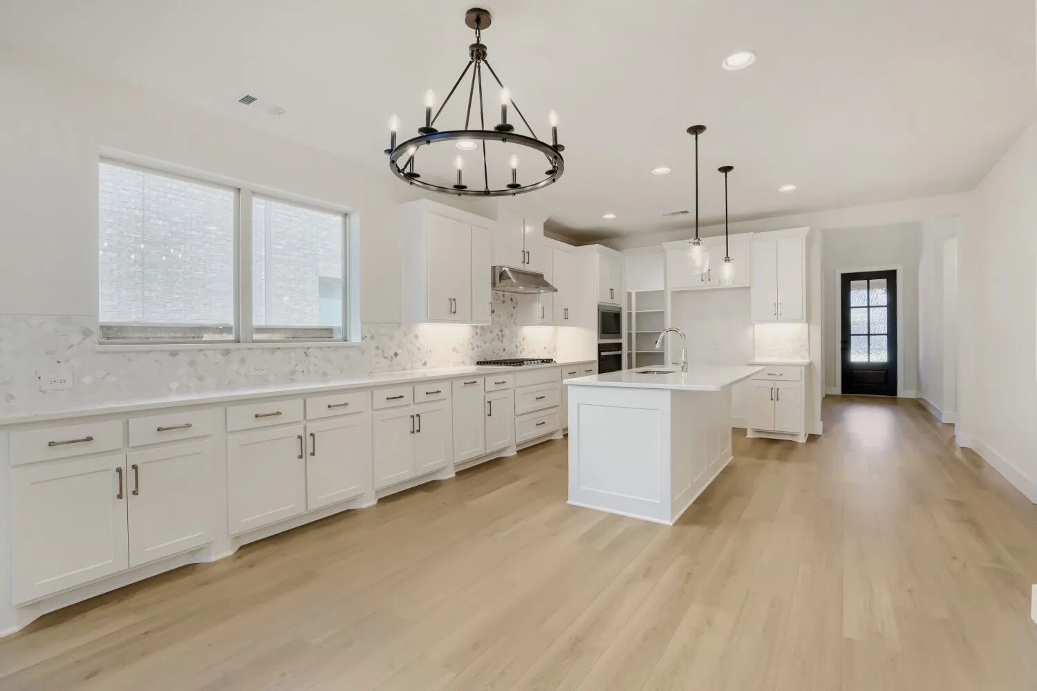 Open-concept kitchen featuring white shaker cabinetry, an expansive central island with a sink, stainless steel appliances, and wood-finish flooring