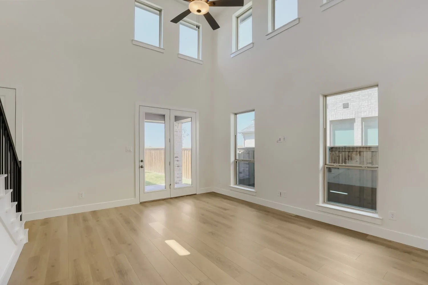 Two-story ceiling, wood-finish flooring, multiple windows, double-door exterior access, and a ceiling fan with light fixture