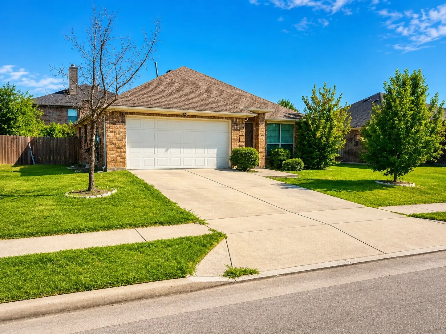 Charming brick home featuring a two-car garage with white paneled doors, situated on a manicured lawn with mature landscaping including evergreen shrubs and shade trees