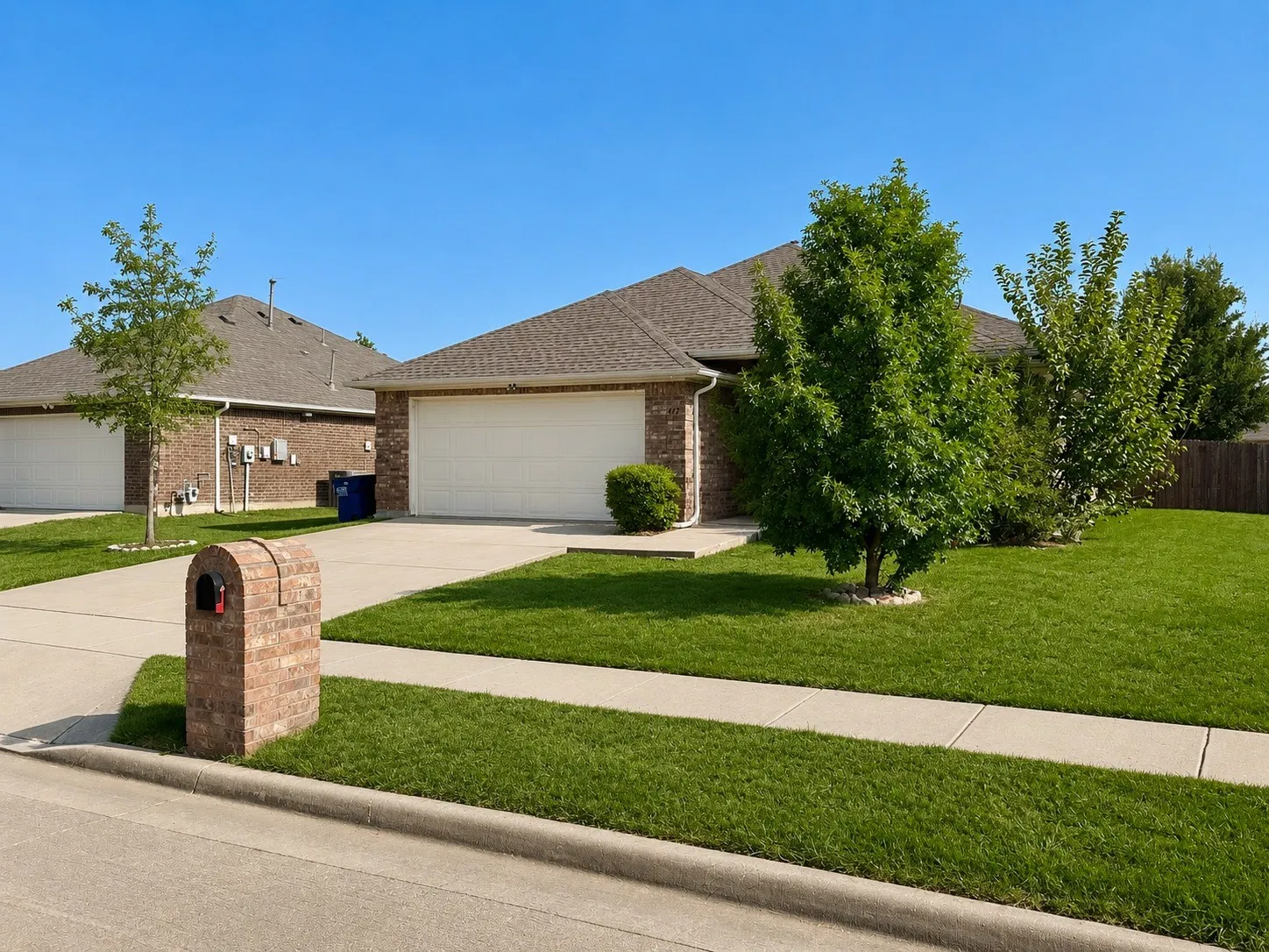 Brick exterior home featuring a spacious driveway, two-car garage, and manicured front lawn with mature landscaping including established trees and shrubs