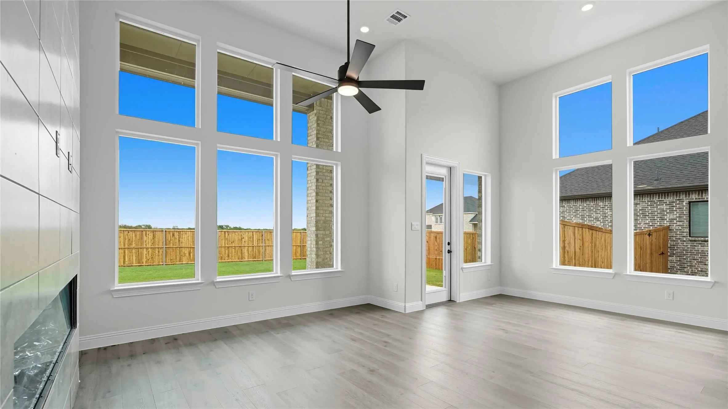 Bright and spacious interior showcasing abundant natural light through multiple large multi-pane windows, wide-plank wood-finish flooring, ceiling fan, and clean white walls