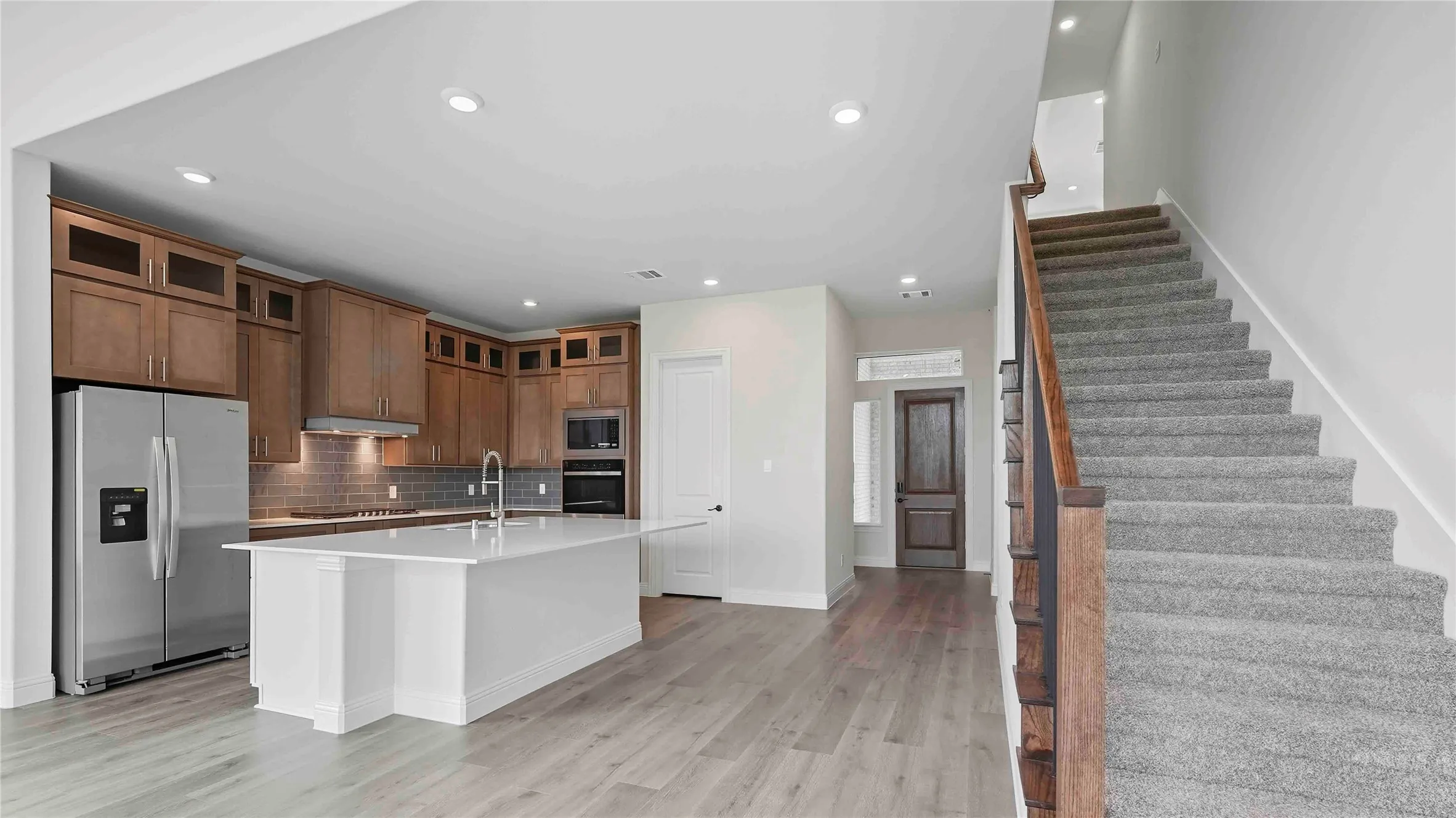 Open-concept kitchen featuring wood-finish cabinetry, stainless steel appliances, a center island with a sink, and light wood-finish flooring extending into the entry hall
