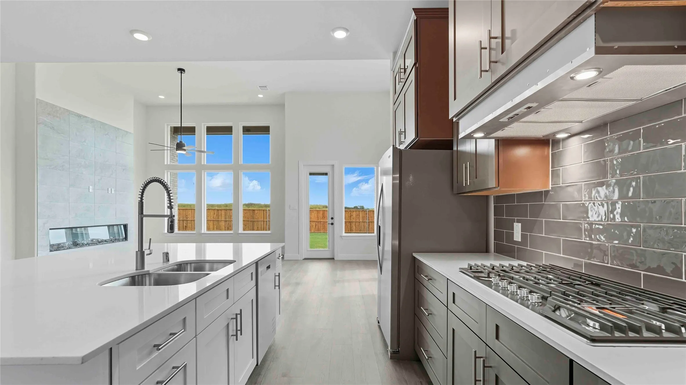 Galley-style kitchen featuring white quartz countertops, gray cabinetry with brushed nickel hardware, a six-burner gas cooktop, stainless steel undermount sink with pull-down faucet, subway tile backsplash, and abundant natural light from multi-pane glass doors and windows overlooking the property
