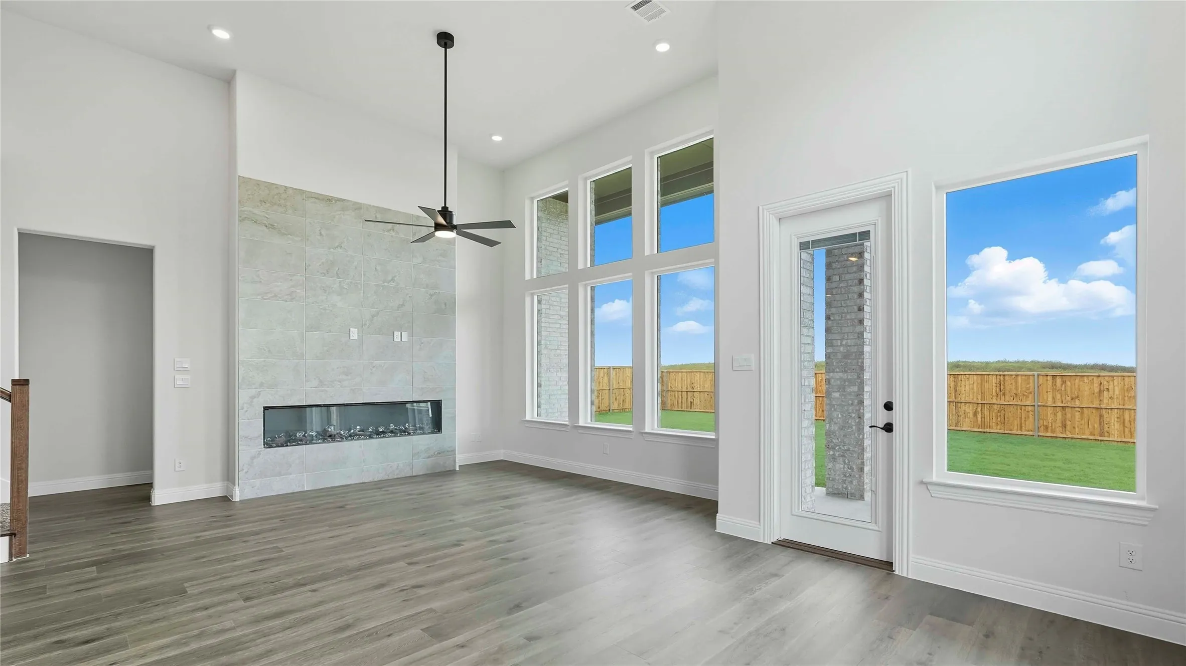 Living space featuring high ceilings, wood-finish flooring, a wall-mounted linear fireplace with a textured tile surround, a ceiling fan, and multiple large windows overlooking a fenced backyard