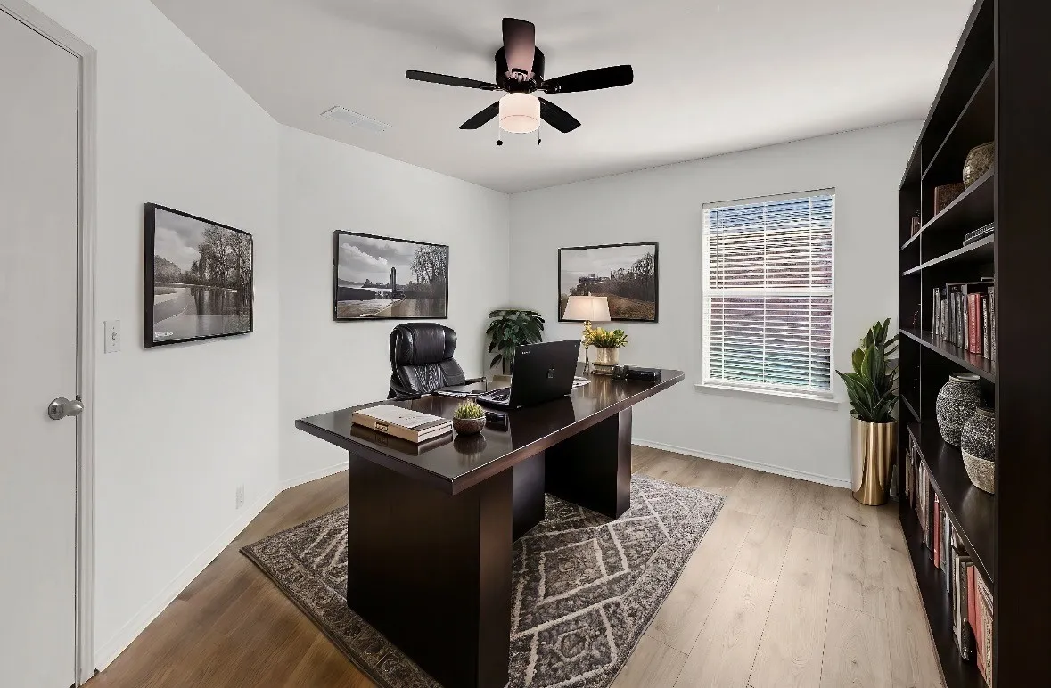 Sophisticated home office featuring a dark wood executive desk, built-in shelving with display storage, hardwood flooring, geometric area rug, black ceiling fan with integrated lighting, framed architectural artwork, potted greenery, and abundant natural light from a large window