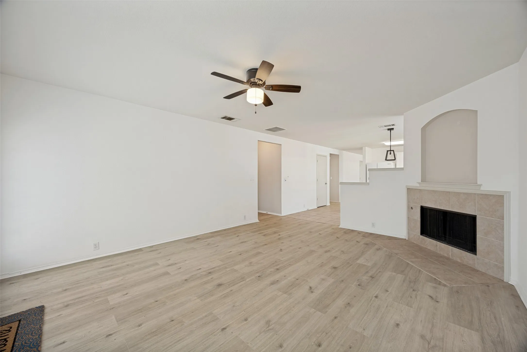 Spacious living area featuring light wood-finish flooring, a ceiling fan with integrated lighting, and a corner fireplace with a tiled surround and mantel