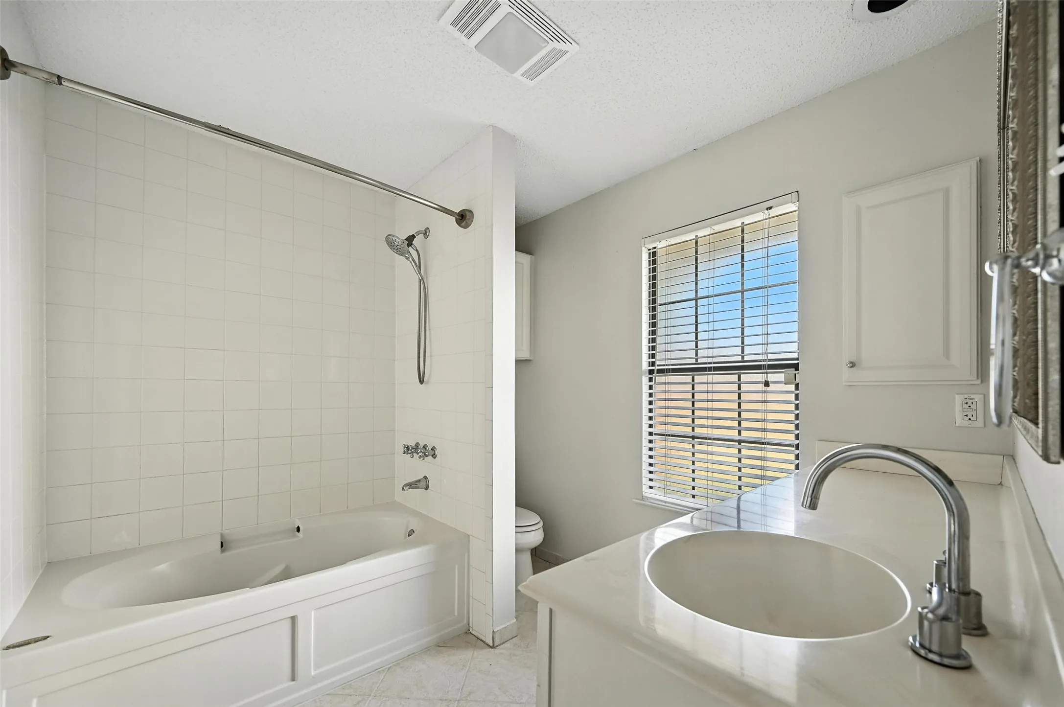 Bathroom featuring a white tiled tub and shower combination with a handheld showerhead, a single vanity with an integrated sink and chrome faucet, and a window with horizontal blinds