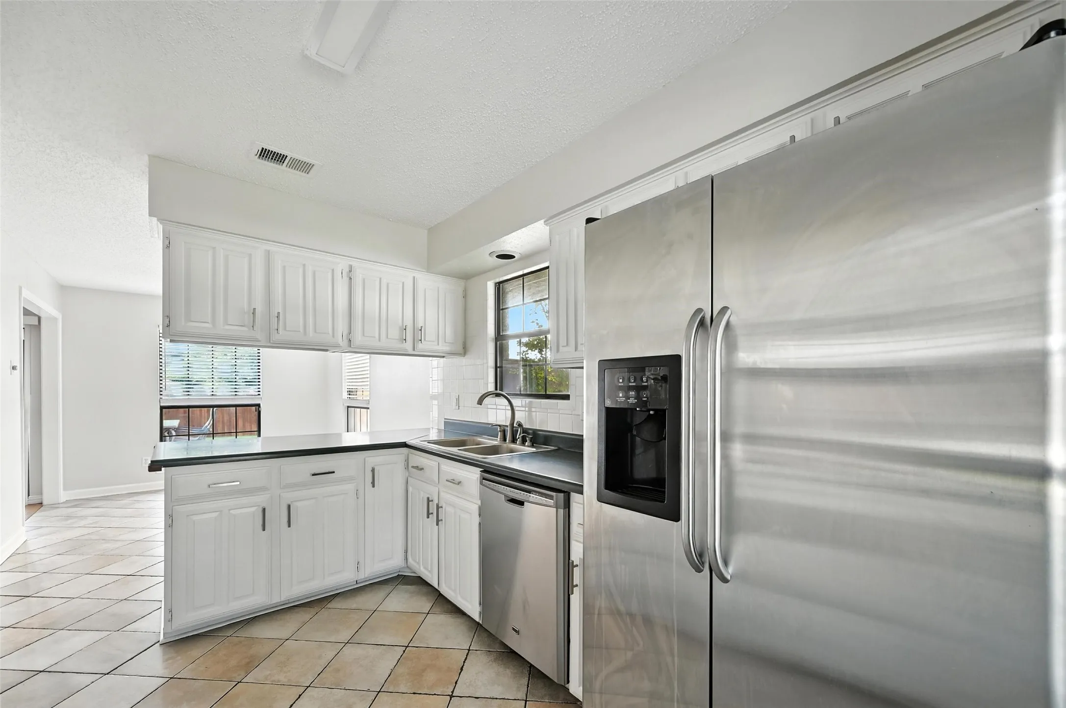 Bright, open kitchen featuring crisp white cabinetry with ample storage, stainless steel appliances including a side-by-side refrigerator and dishwasher, black countertops, a functional island with prep space, and ceramic tile flooring throughout