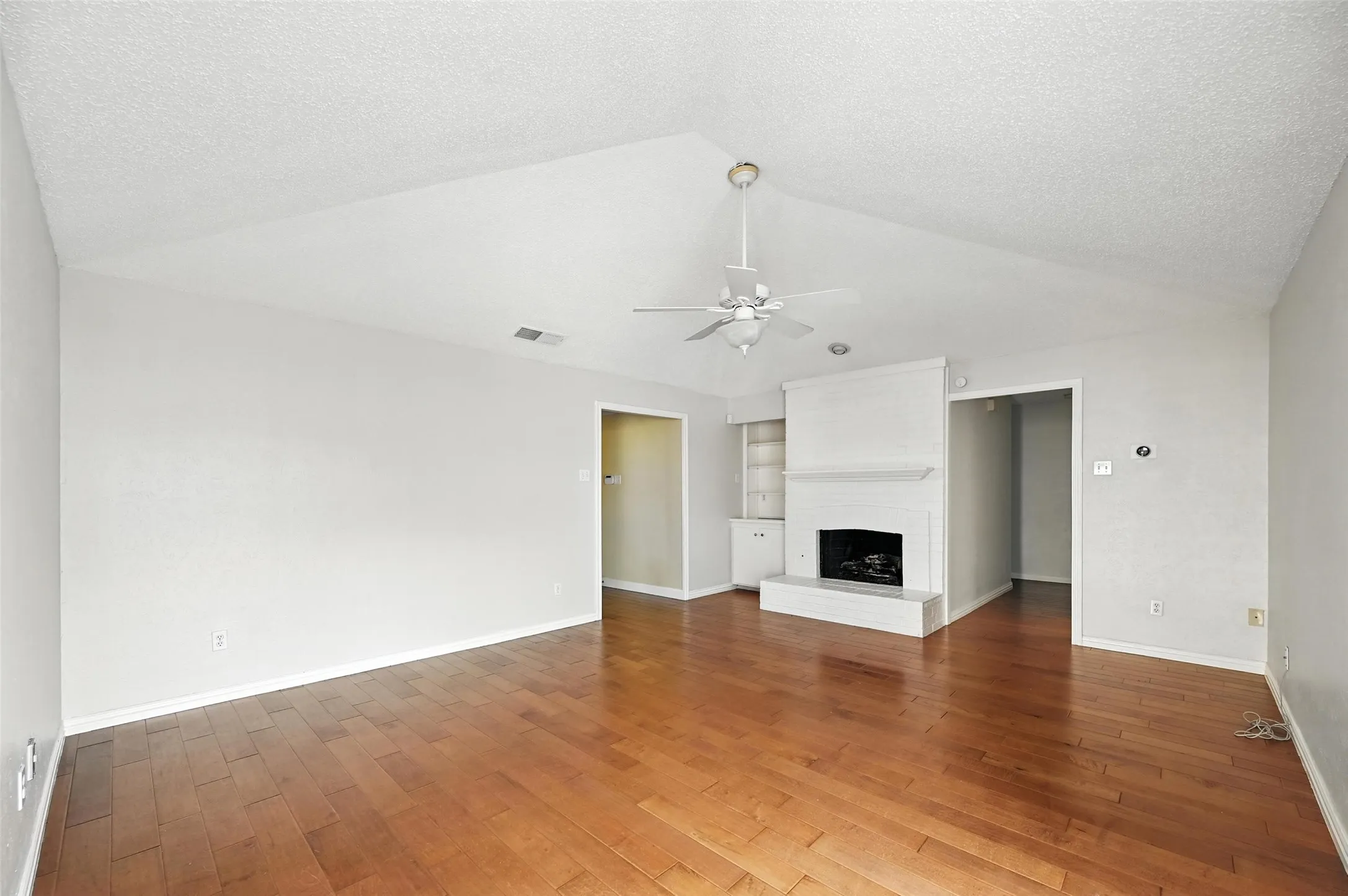 Spacious open-concept living area featuring wood-finish flooring, vaulted ceilings with ceiling fan, and a gas fireplace with built-in white cabinetry