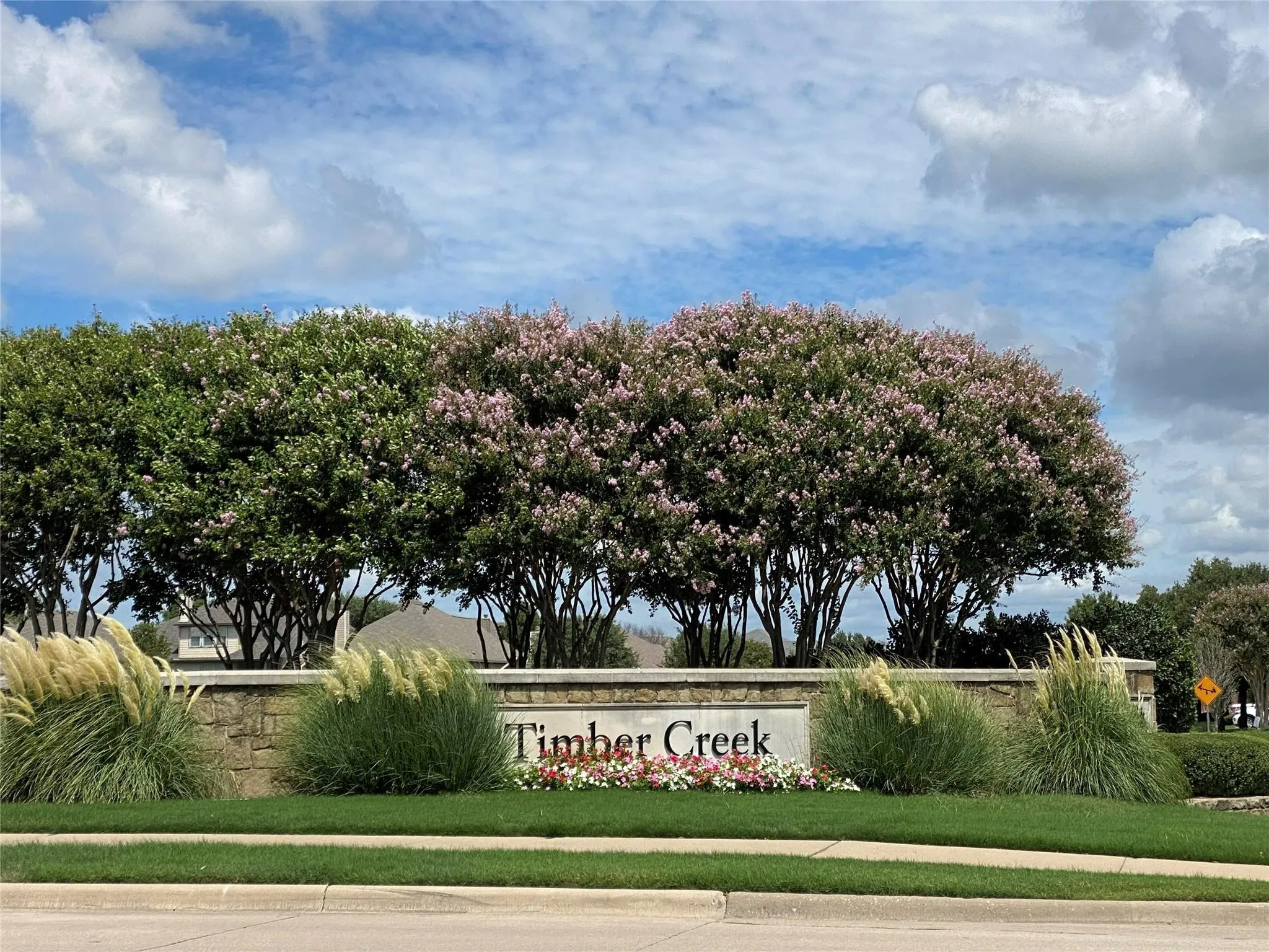 Stone entry monument featuring "Timber Creek" lettering, framed by flowering trees and ornamental grasses