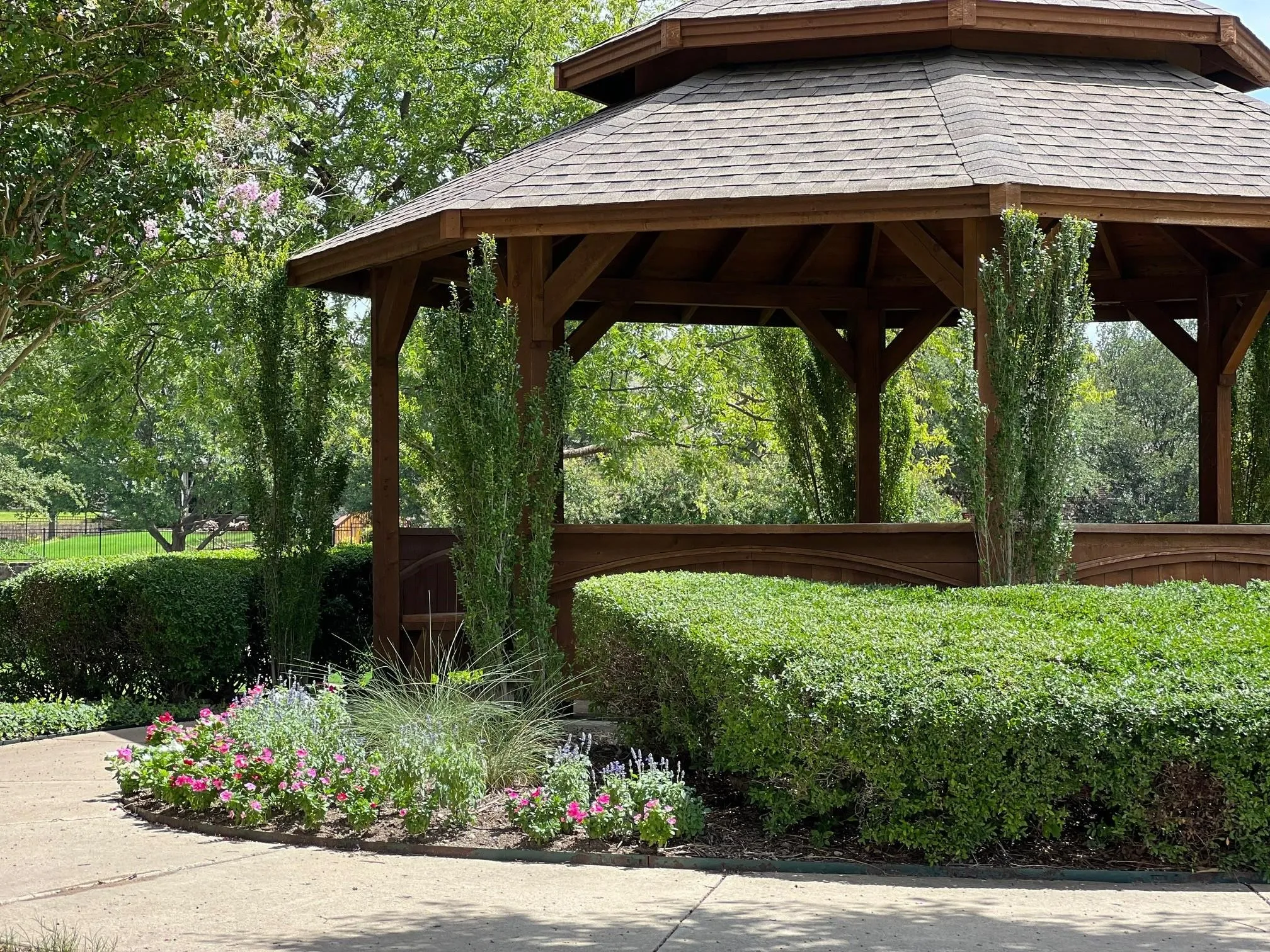 Wooden gazebo with a shingled roof, set amidst mature trees and meticulously maintained hedges