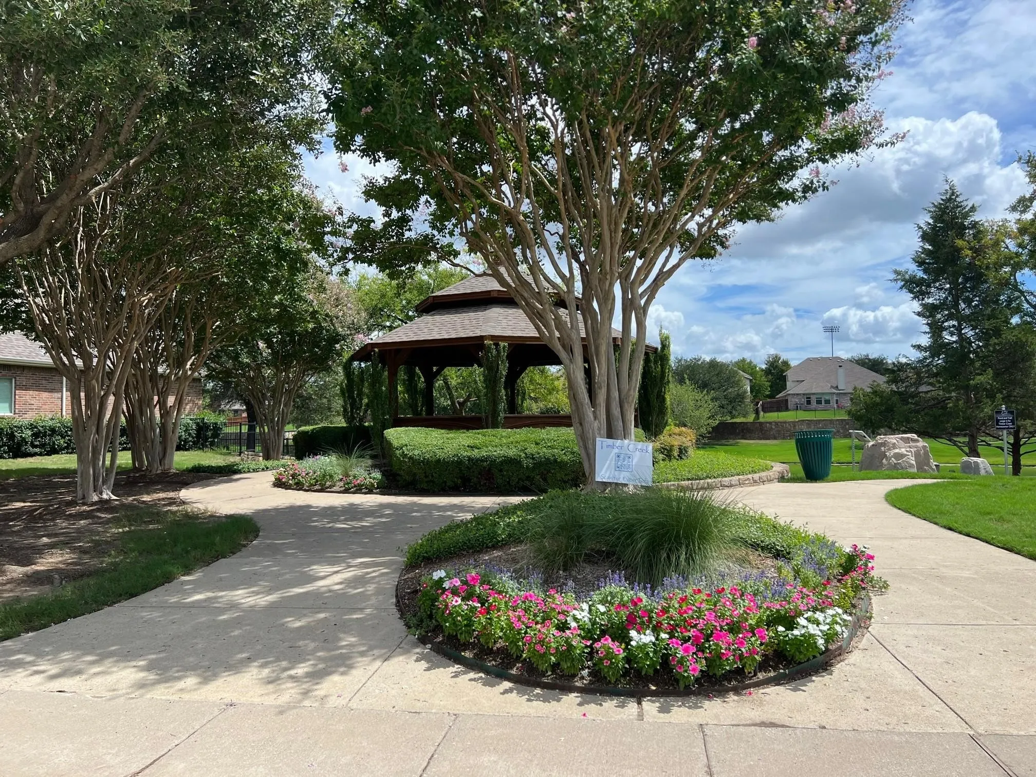 Central gazebo structure with a hip roof, surrounded by mature trees, manicured hedges, and a circular planting bed featuring colorful flowers and ornamental grasses