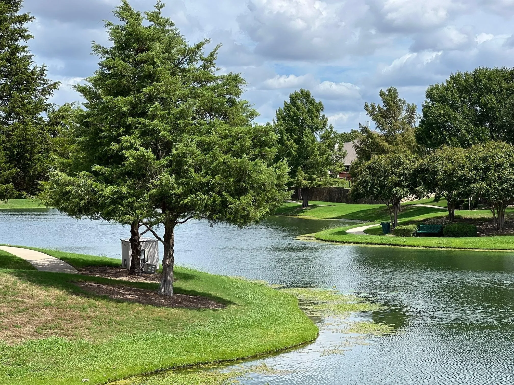 Lush green landscape featuring a serene water body, mature trees, manicured lawns, and paved pathways