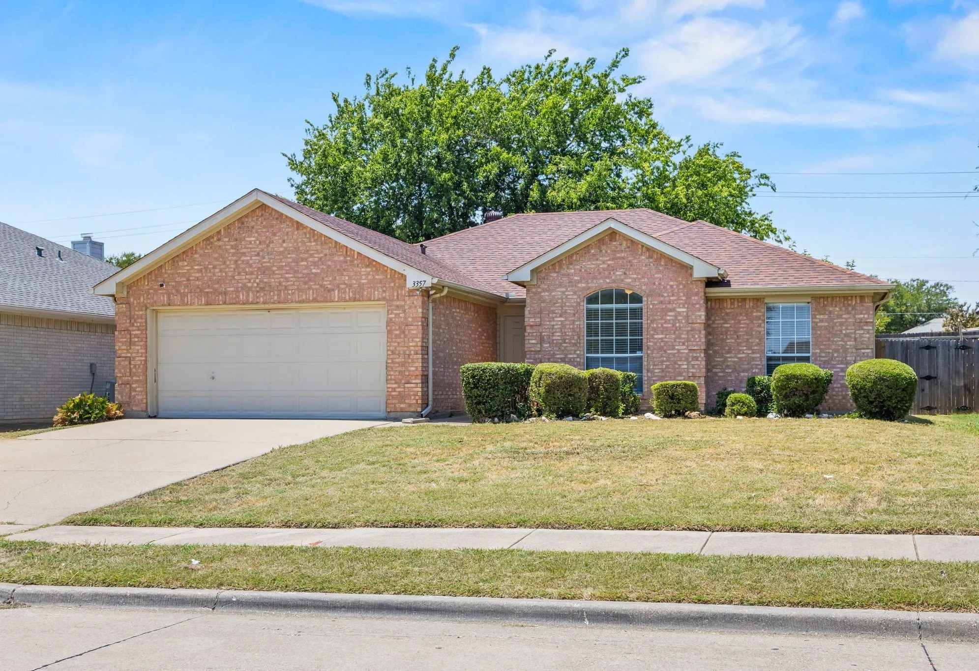 Brick exterior featuring a two-car garage, mature landscaping, an arched window, and a fenced backyard