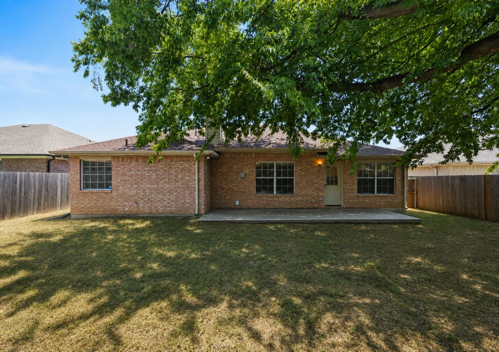 Brick single-story home featuring a spacious rear patio with mature shade trees, manicured lawn, and wooden privacy fencing