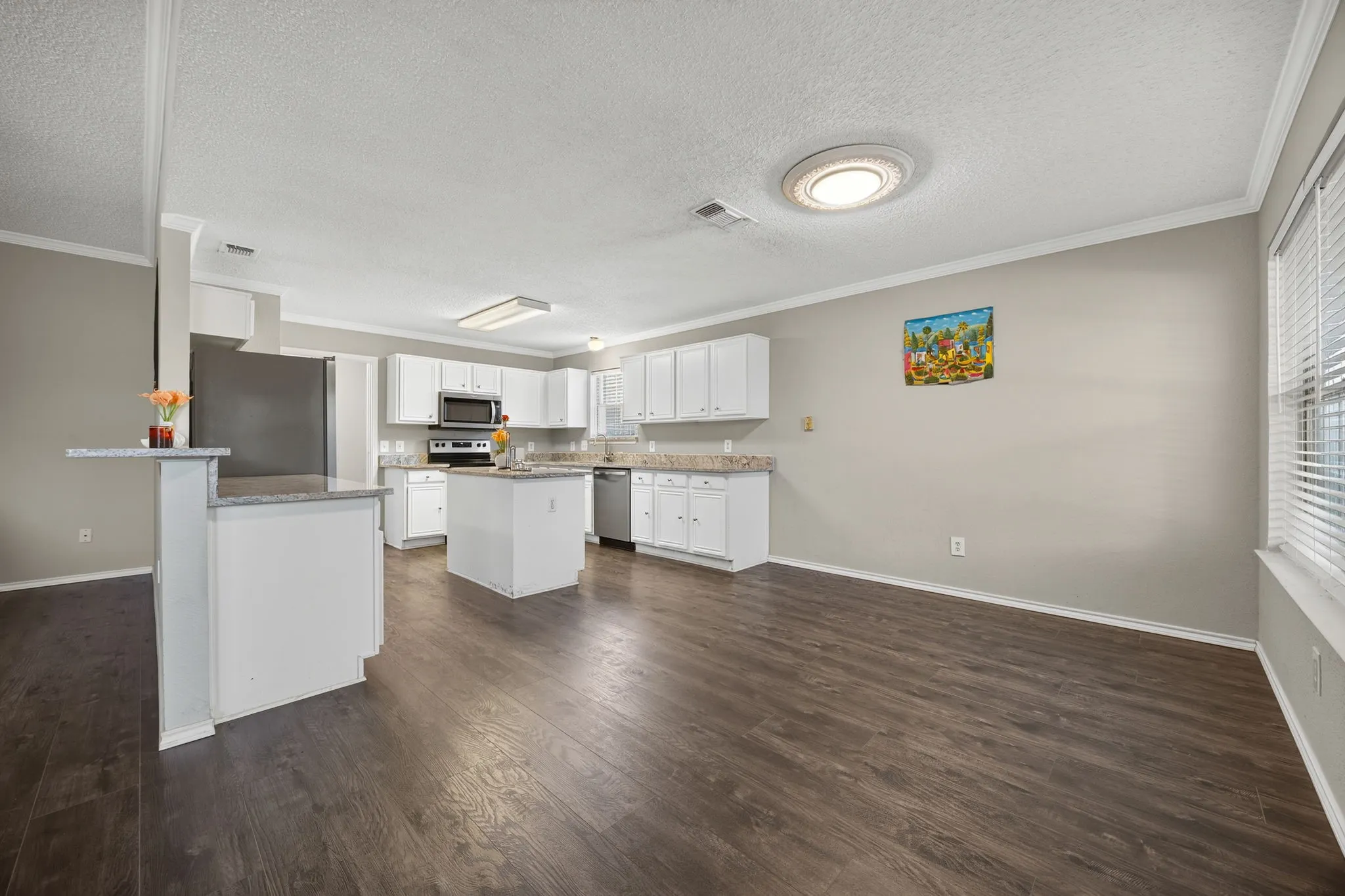 Open-concept kitchen and living area featuring white cabinetry, stainless steel appliances, granite countertops, a large island with seating, and wood-finish flooring throughout