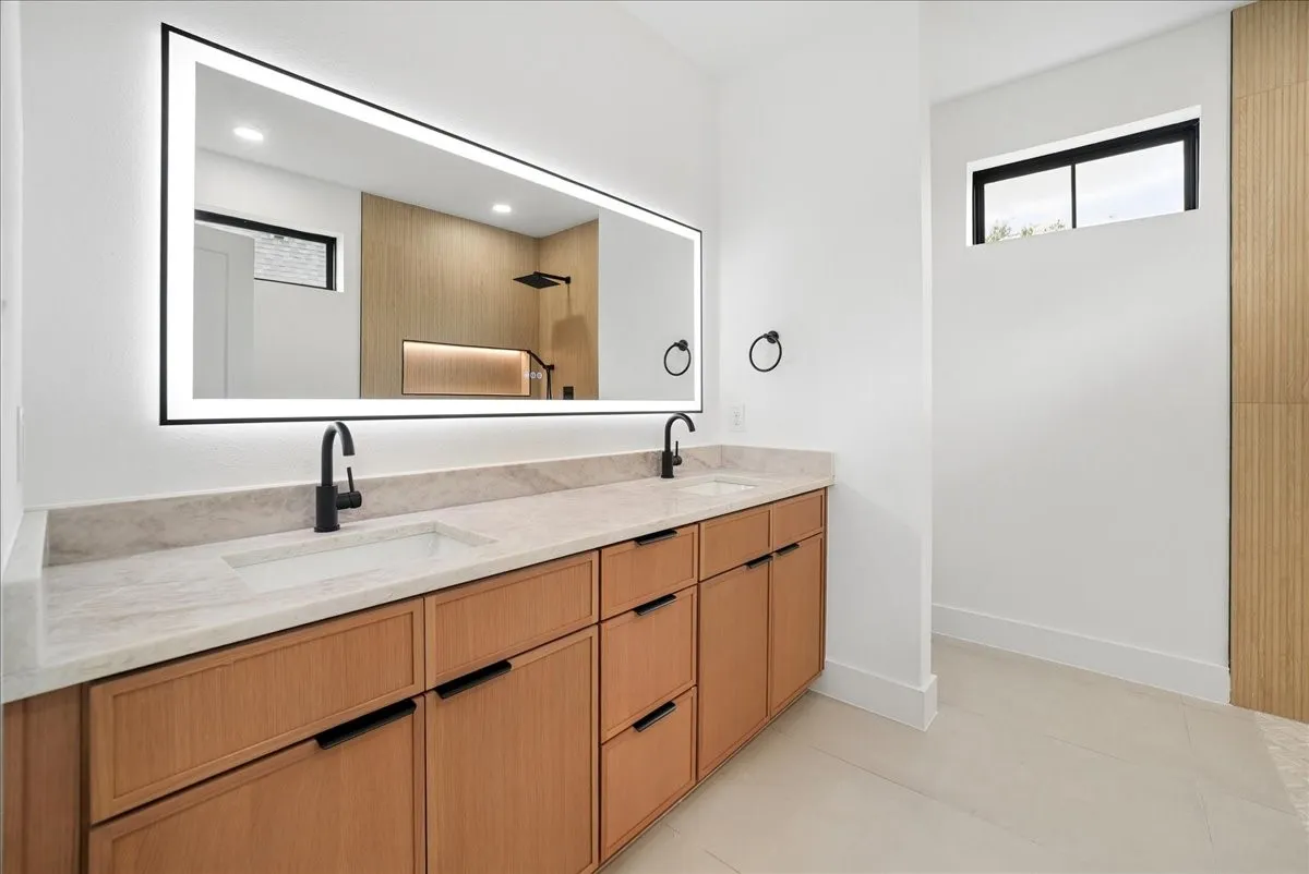 Dual vanity with undermount sinks and matte black faucets, integrated illuminated mirror, light-toned wood-finish cabinetry, and a stone countertop