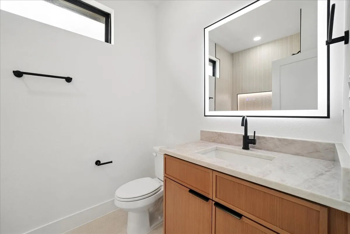 Illuminated vanity mirror, wood-finish cabinetry, integrated sink, stone countertop, and matte black fixtures
