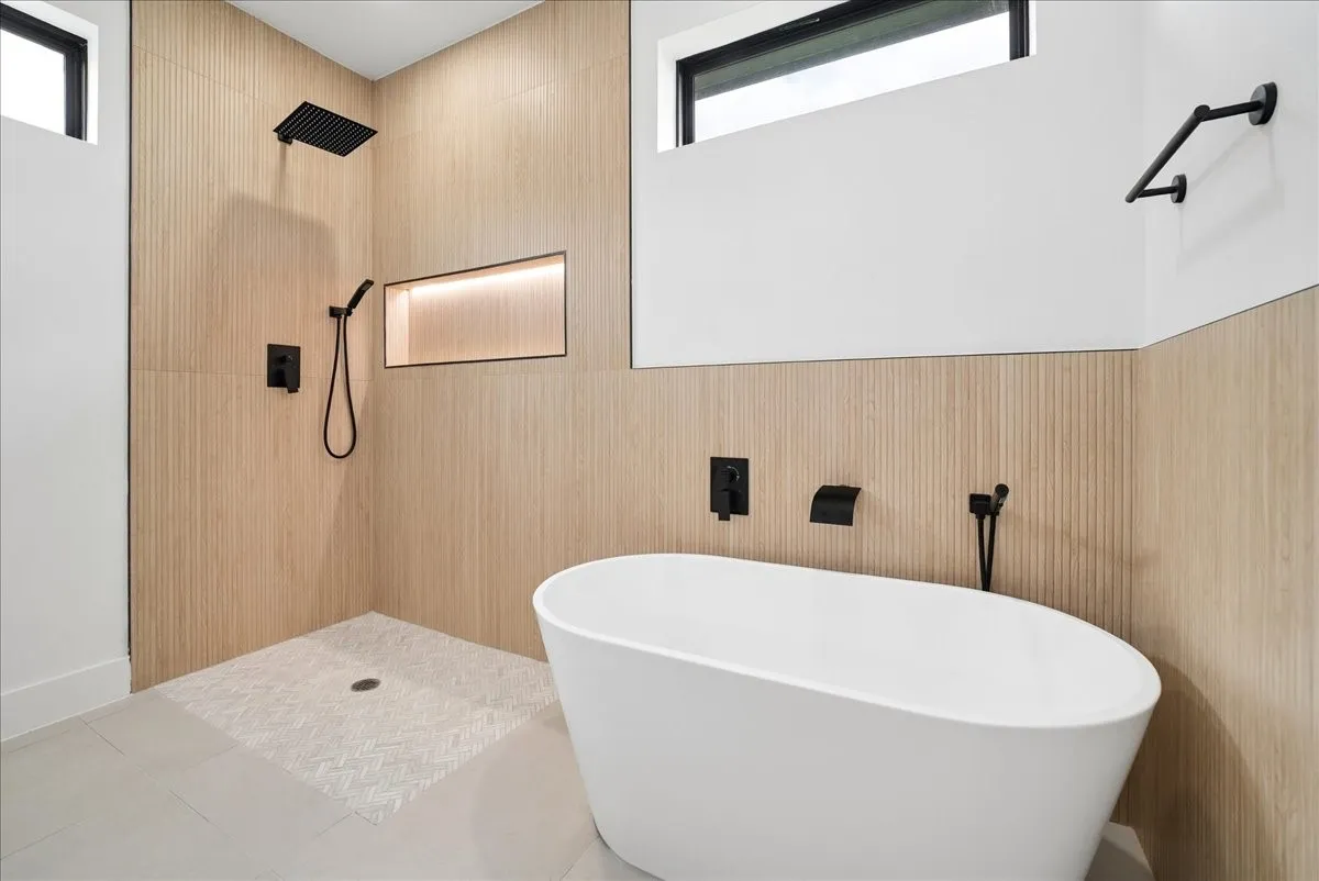 Freestanding soaking tub with matte black fixtures, an illuminated shower niche, a rain showerhead, and light wood-finish wall paneling